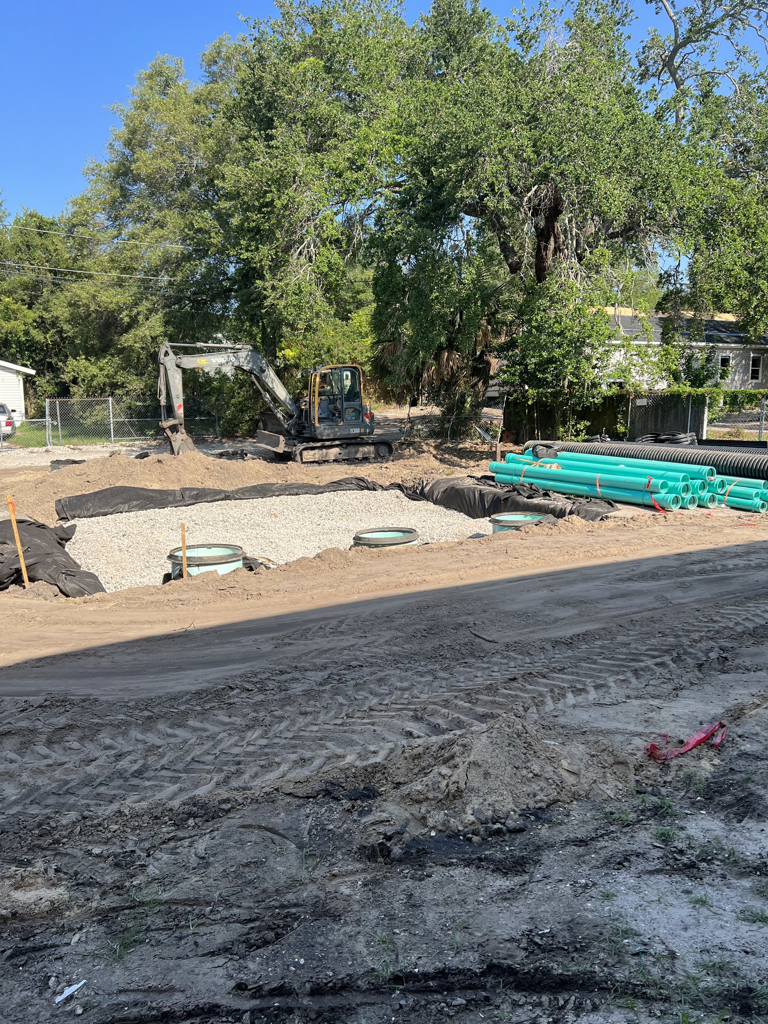 Construction site with exposed soil and gravel, green pipes stacked on the right, a small excavator in the background near a chain-link fence, trees and a house in the distance, under a clear blue sky.