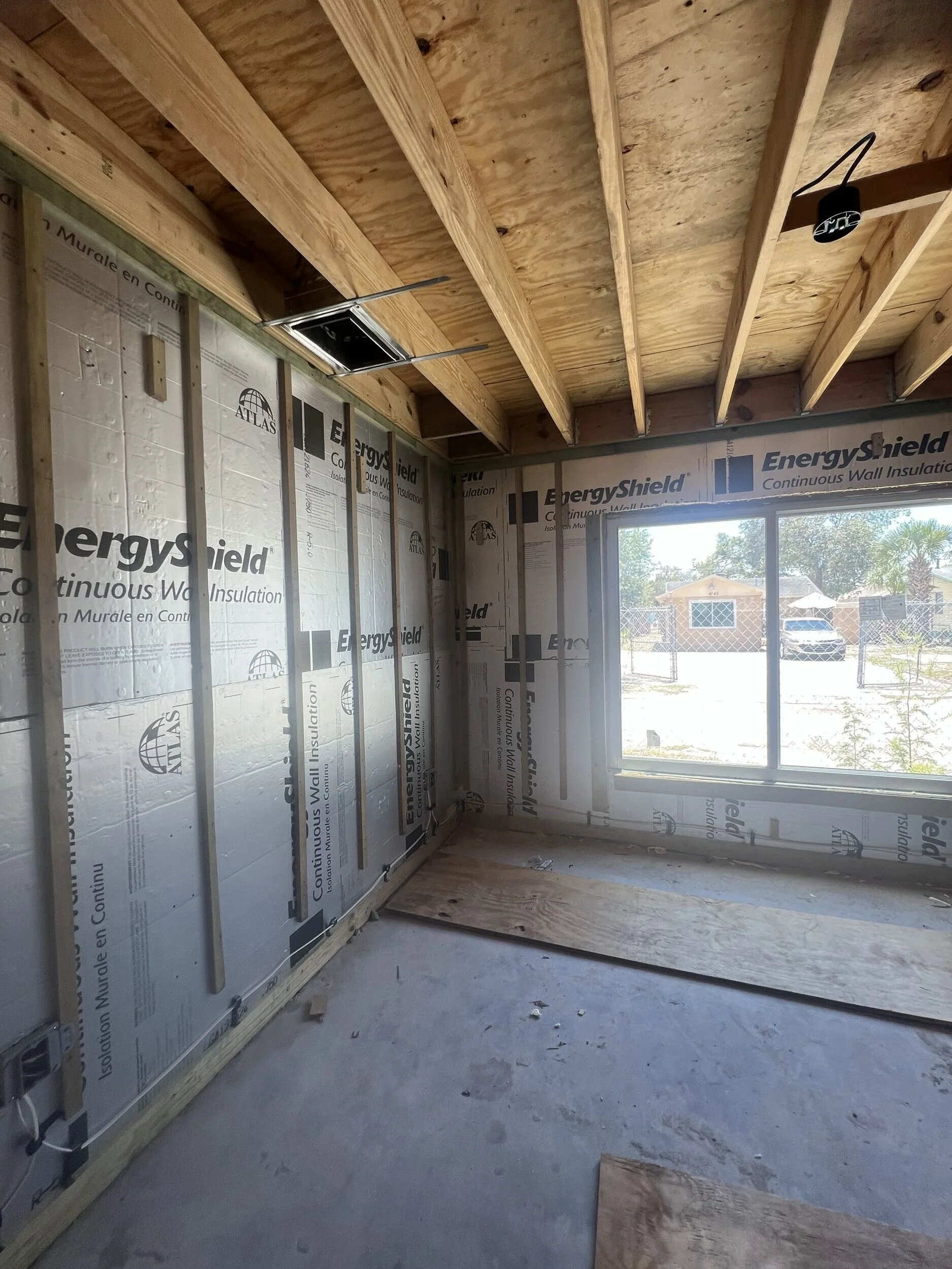 Interior of a room under construction with exposed wooden framing and insulation. There is a window showing an outdoor scene with trees, a house, and a parked car.