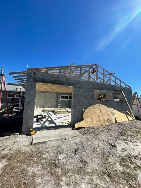 Construction site of a building with concrete block walls and wooden framing on the upper level, showing building materials, tools, and ladders under a clear blue sky.