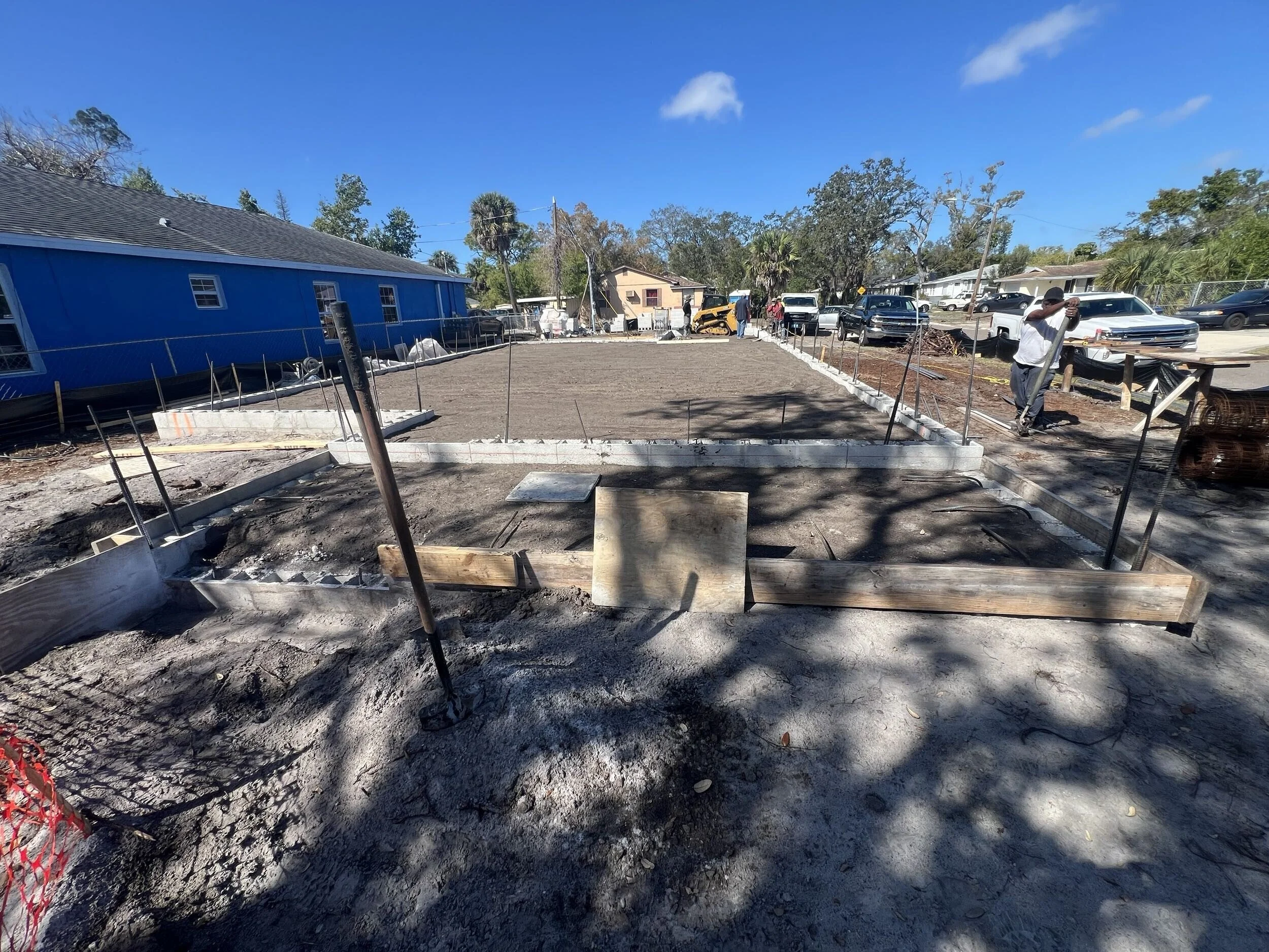 Construction site with an empty foundation, workers, and vehicles, in a residential neighborhood with trees and houses, under a blue sky.