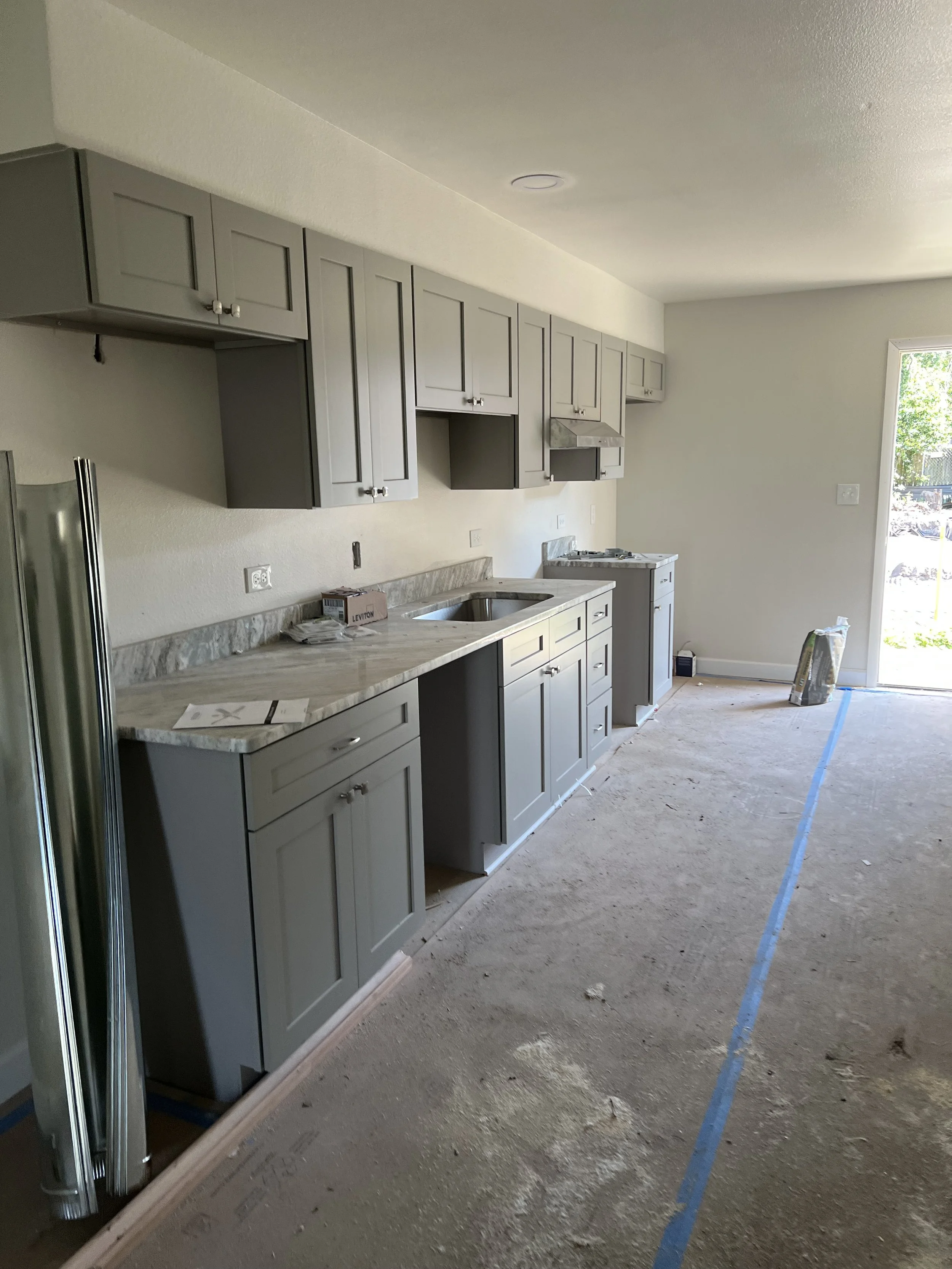 Interior view of a kitchen under construction with gray cabinets, a marble countertop, and some construction materials and tools on the floor.