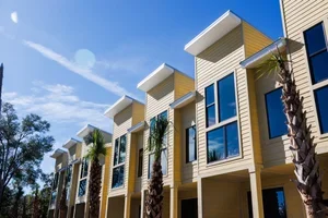 Row of modern townhouses with yellow siding, large windows, and small front porches, featuring palm trees in front on a sunny day.