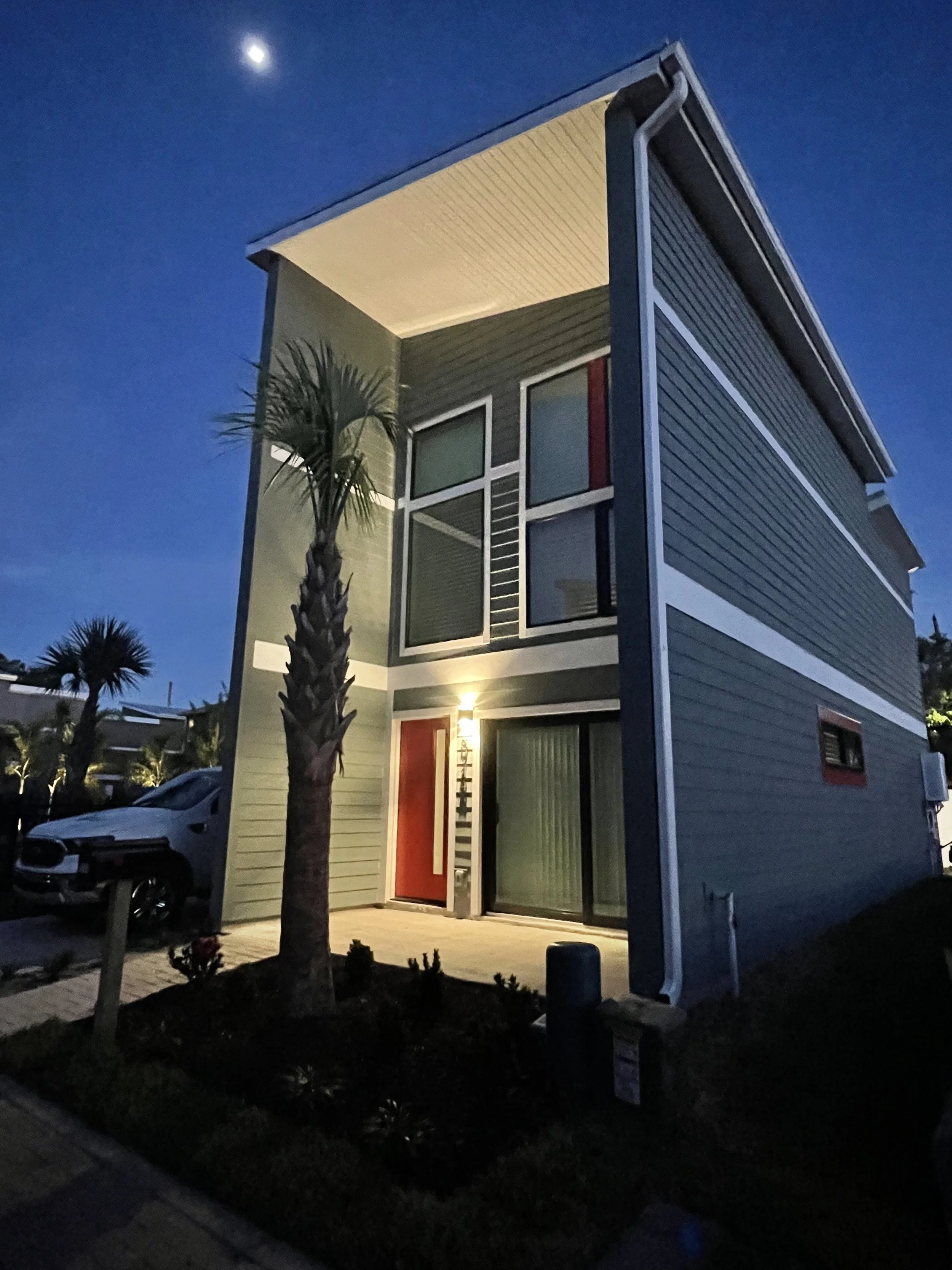 A modern, multi-story house with a dark exterior, large windows, and a red front door at night. The house has a sloped roof and a small landscaped area with a palm tree in the front yard. The moon is visible in the sky.