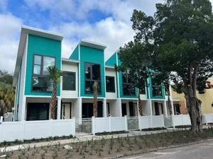 Modern multi-story residential building with blue and white exterior, palm trees in front, and a white fence, under a partly cloudy sky.