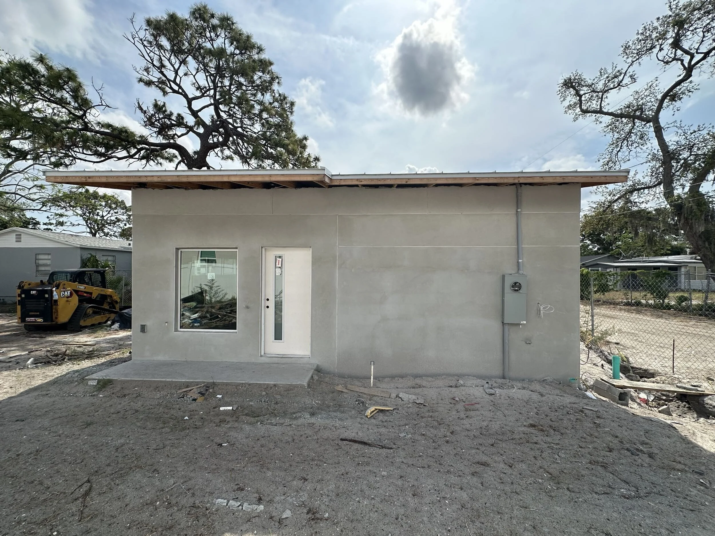 A partly constructed building with a flat roof, a door and window, situated on a dirt lot. Construction equipment and debris are visible nearby, with trees and blue sky with clouds in the background.