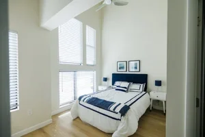 Bedroom with white walls, large windows with blinds, a bed with blue and white bedding, and two bedside tables with lamps