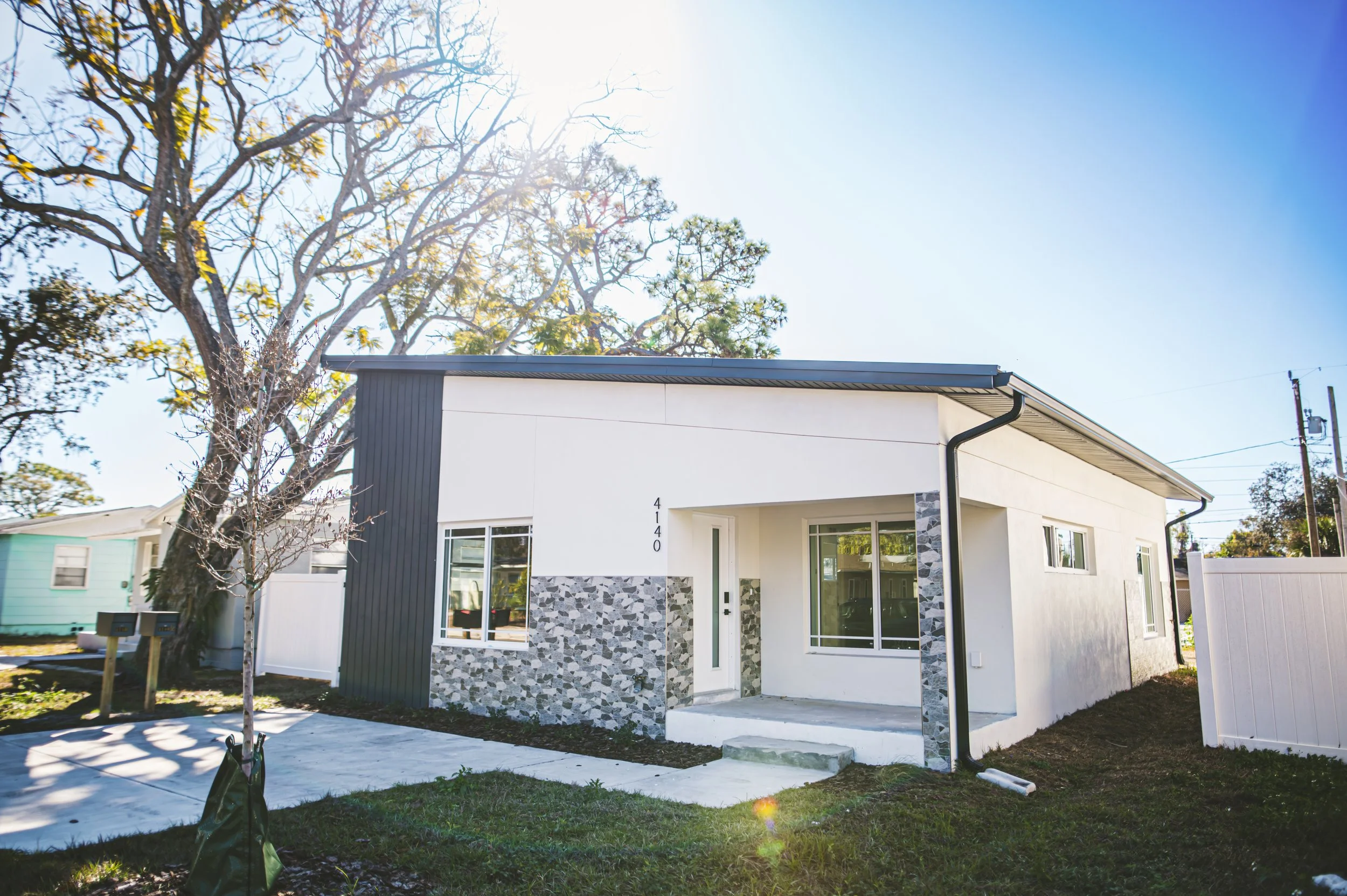 Modern single-story house with white exterior walls, a decorative stone accent wall, large windows, and a dark-colored roof. Front yard with a small tree and a concrete pathway leading to the entrance. Bright sunlight over the scene.
