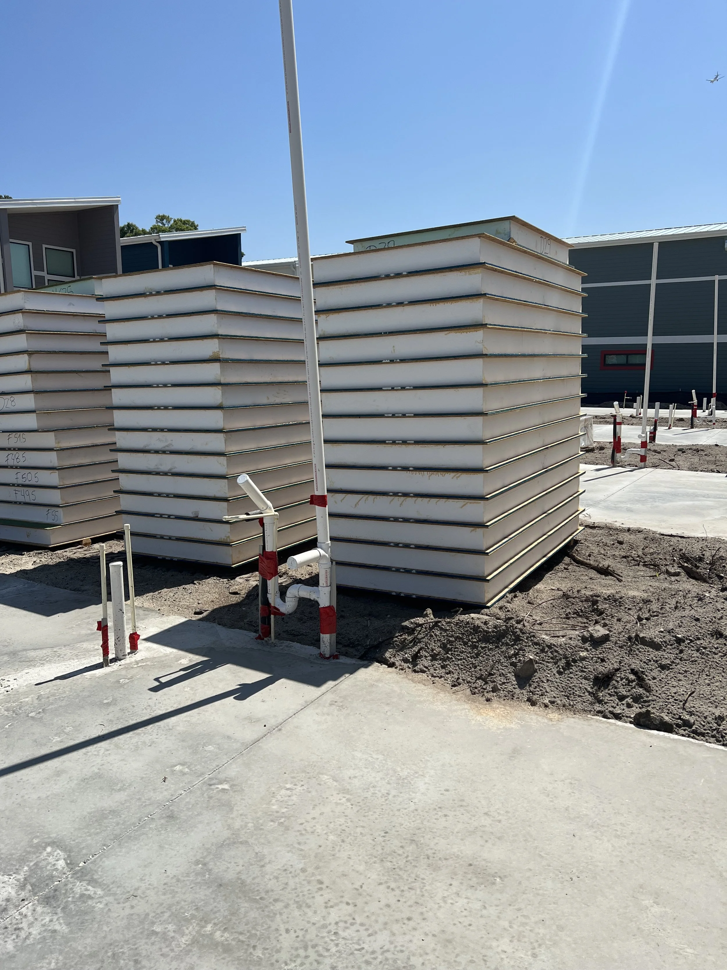 Construction site with large stacked rectangular building materials, a sidewalk, and some construction pipes and barriers, under clear blue sky.