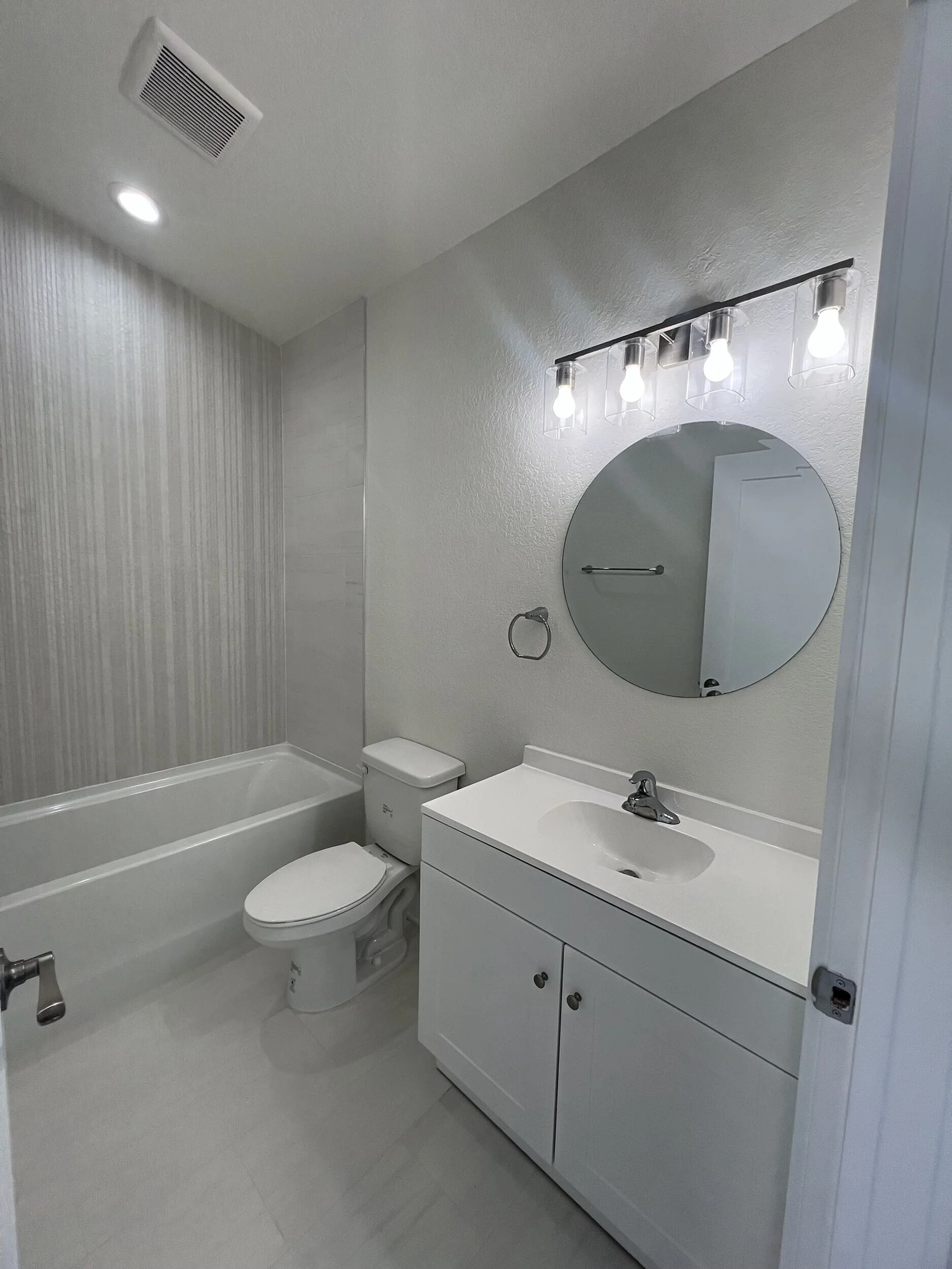 A modern bathroom with a bathtub, toilet, and white vanity with a sink. There are four exposed light bulbs above a round mirror on the wall, and a towel ring beside the mirror. The walls are painted in light, neutral tones, and the ceiling has a vent and recessed lighting.