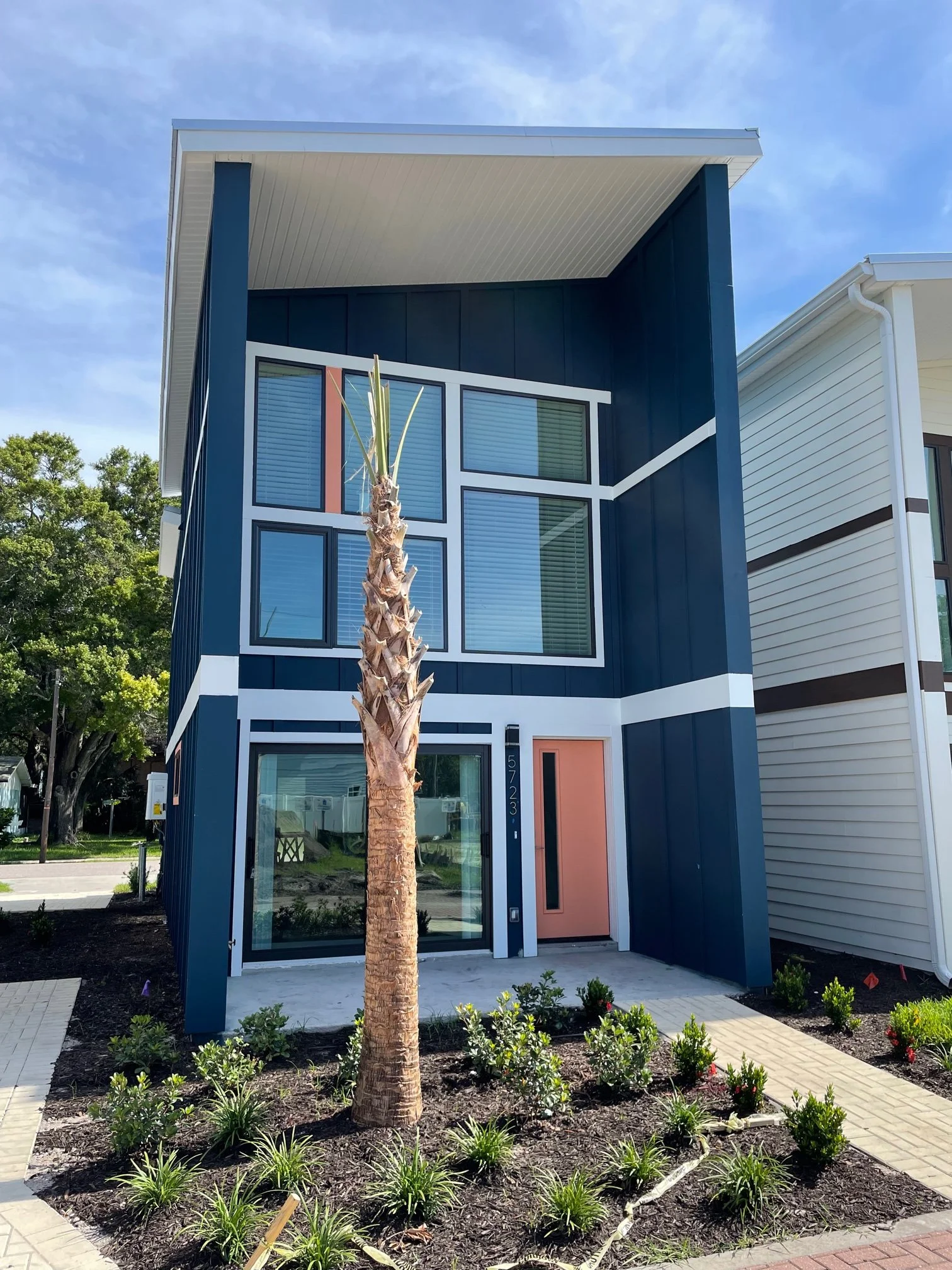 A modern, blue-colored house with a pink front door, large glass windows, and a small front garden with a young palm tree and various plants. The house has a white flat roof and trim, with a clear blue sky in the background.