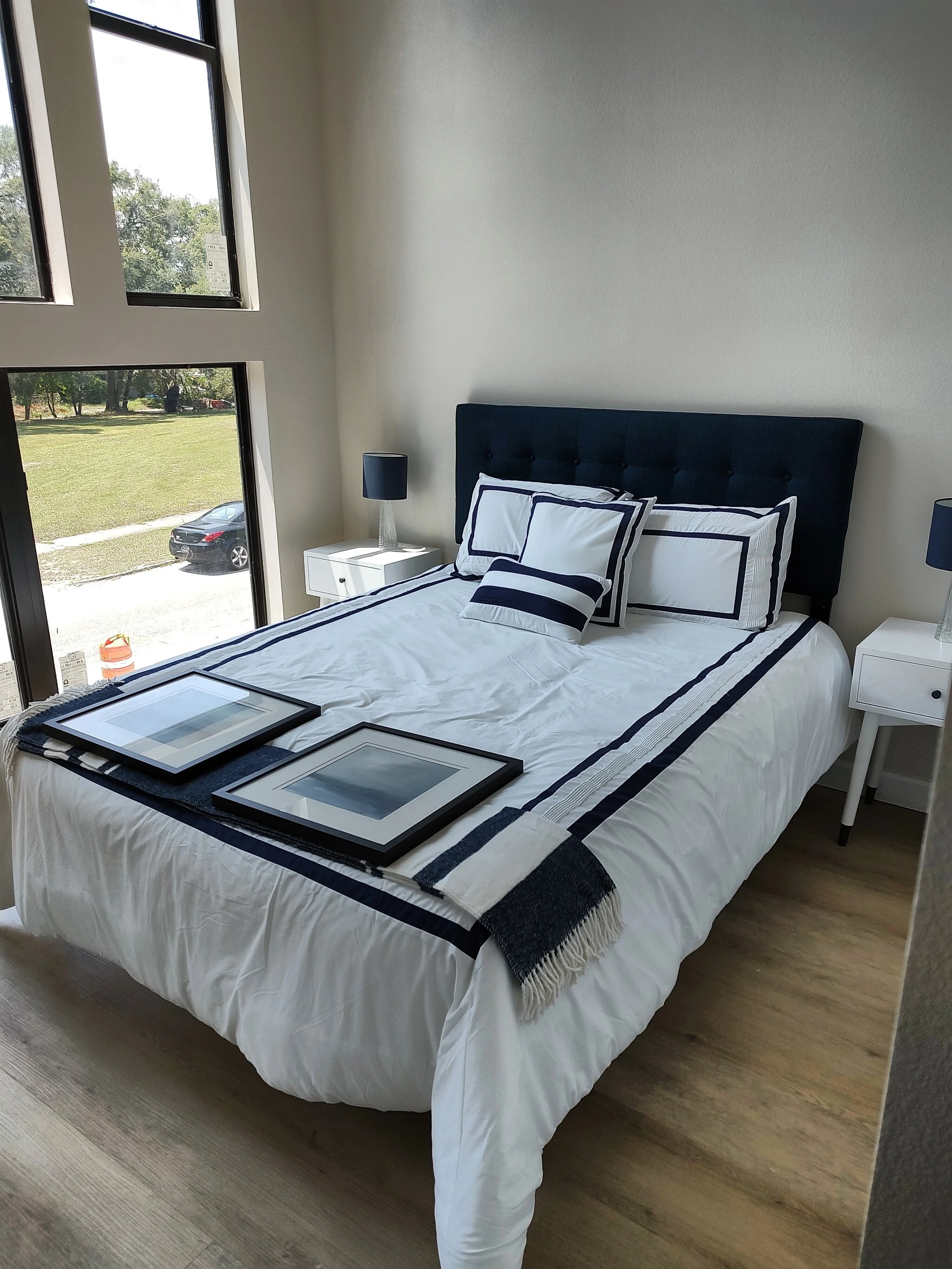A modern bedroom with a black upholstered headboard, white bedding with navy blue trim, and two matching white nightstands with black lamps. The room has large windows letting in natural light and showing a grassy yard outside.