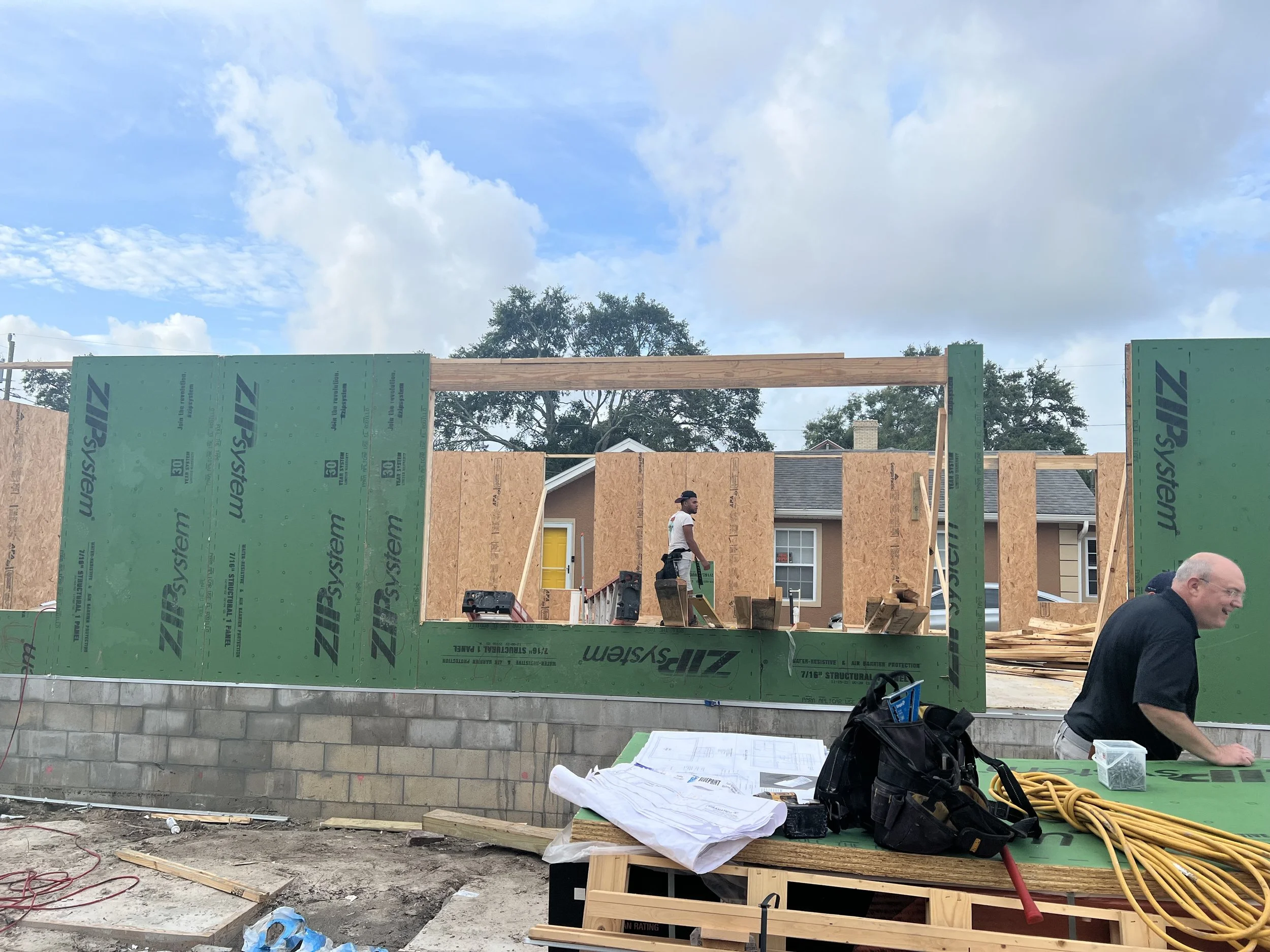 Construction site with framed wall structure, green sheathing, construction tools, and workers under a partly cloudy sky.