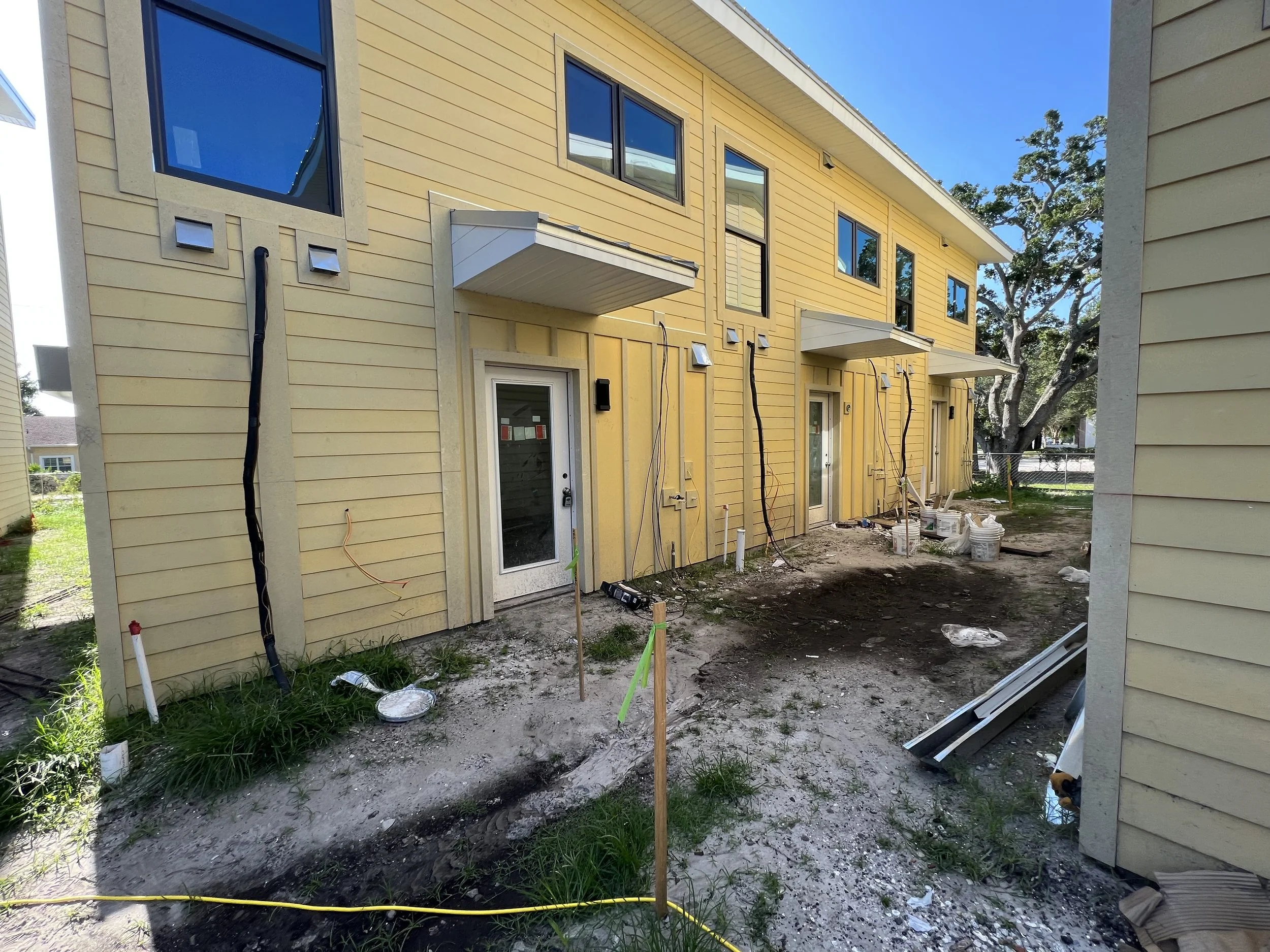 Construction site of a yellow townhouse with several windows, some with black frames. There are construction materials, tools, and pipes on the ground, with some green grass and a tree nearby.