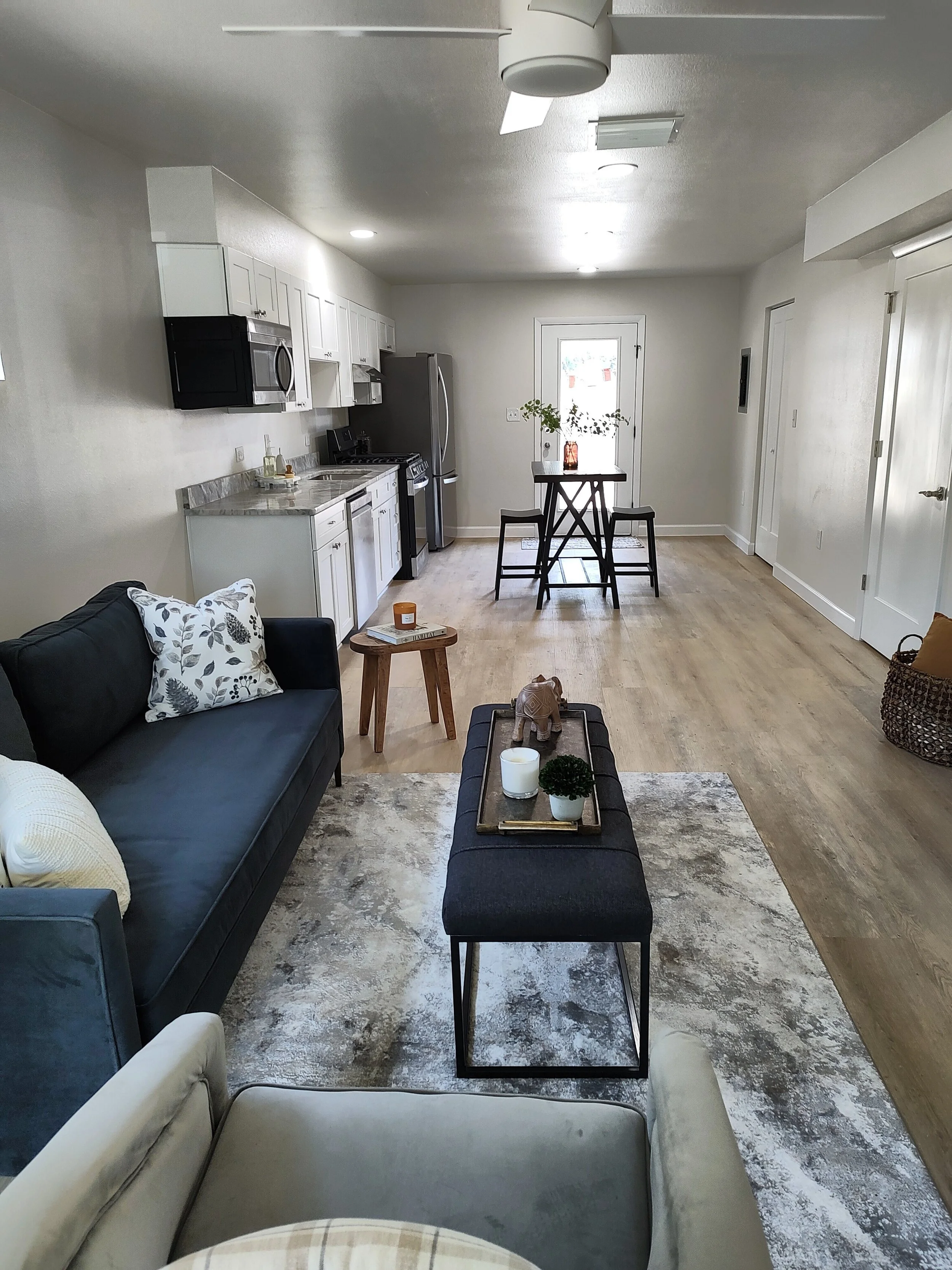 Living room with beige and dark blue sofas, a round wooden side table, a rectangular black coffee table with candles and a small plant, a patterned area rug, and a kitchen in the background with white cabinets, stainless steel appliances, and a small dining table with two chairs near a door with a window.