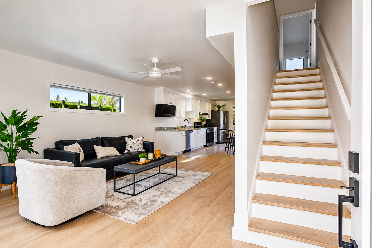 Open-concept living room and kitchen with modern decor, black sofa, white armchair, wooden floors, staircase with white risers and wooden treads, ceiling fan, and a small horizontal window.