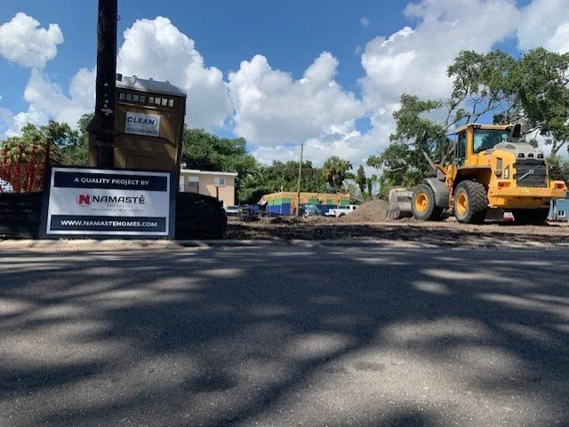 Construction site with a yellow bulldozer on freshly paved asphalt, a sign with the logo for Namaste Asphalt, and a portable toilet under a partly cloudy sky.