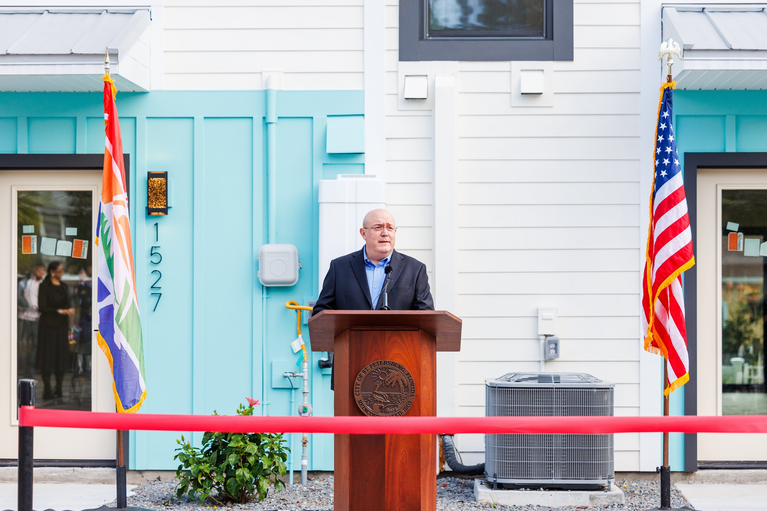 A man in a suit standing behind a podium outdoors, with Flags of the United States and an LGBTQ pride flag on either side, and a building with white siding in the background.