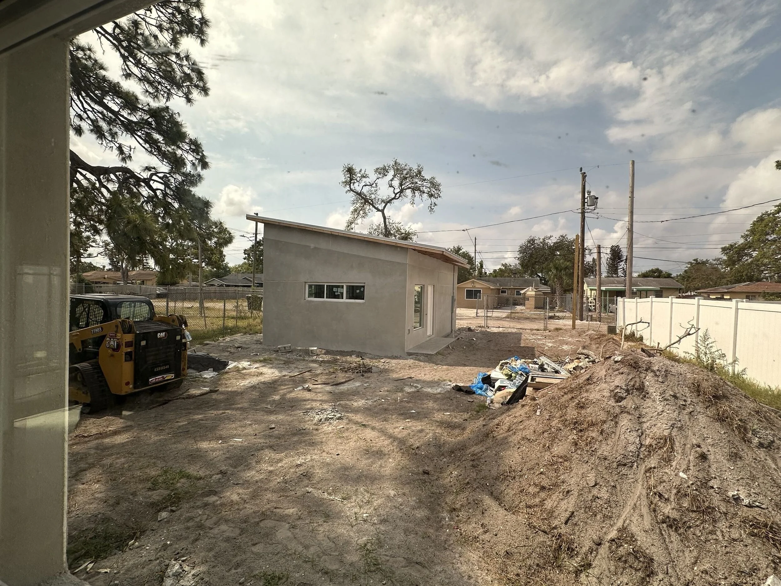 View of a backyard with construction in progress, showing dirt ground, construction tools, a small white building, and a wooden fence. There are power lines and trees in the background, with a partly cloudy sky overhead.