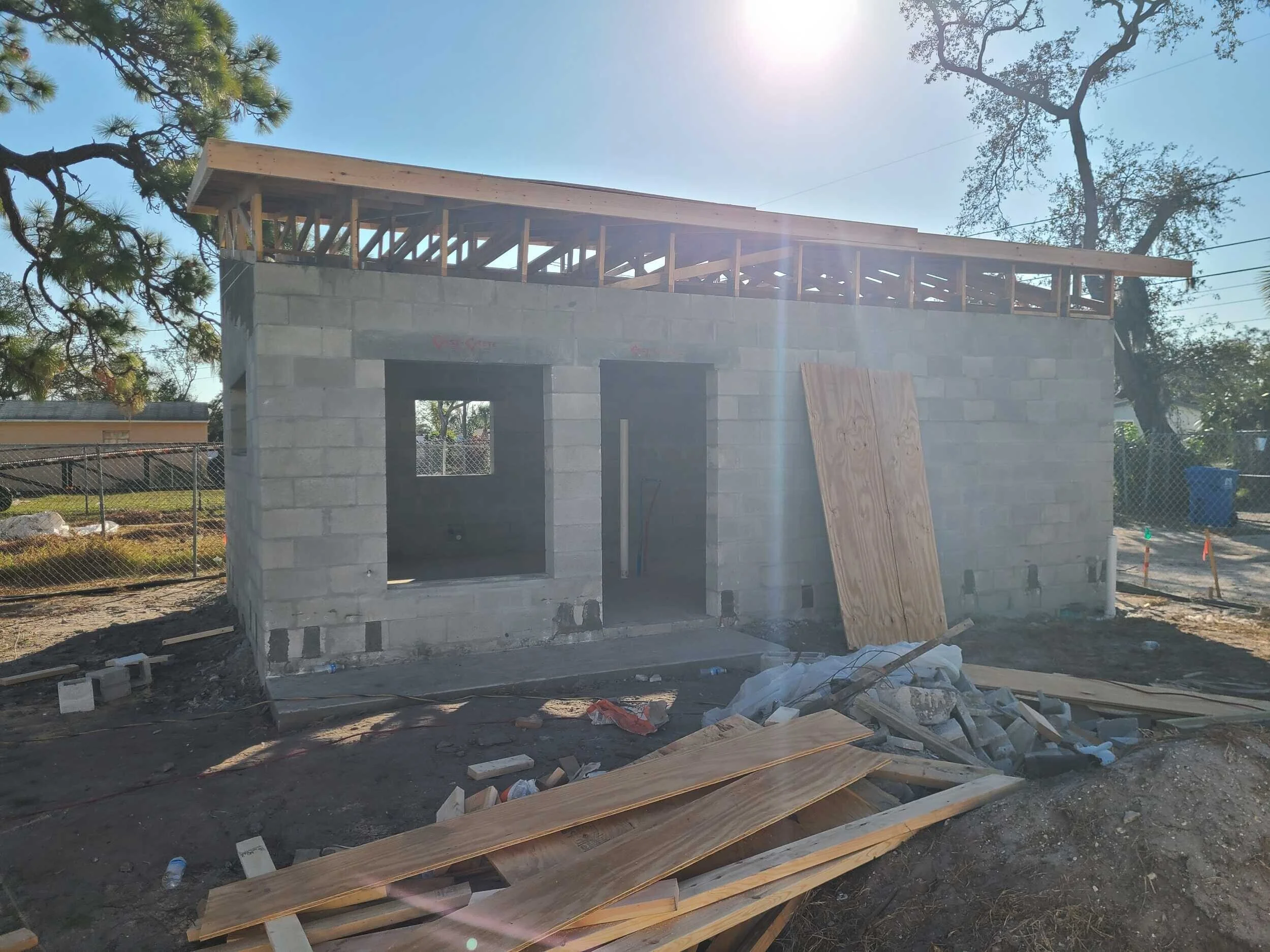 Construction site with a partially built concrete block structure and a wooden roof framework in bright sunlight.