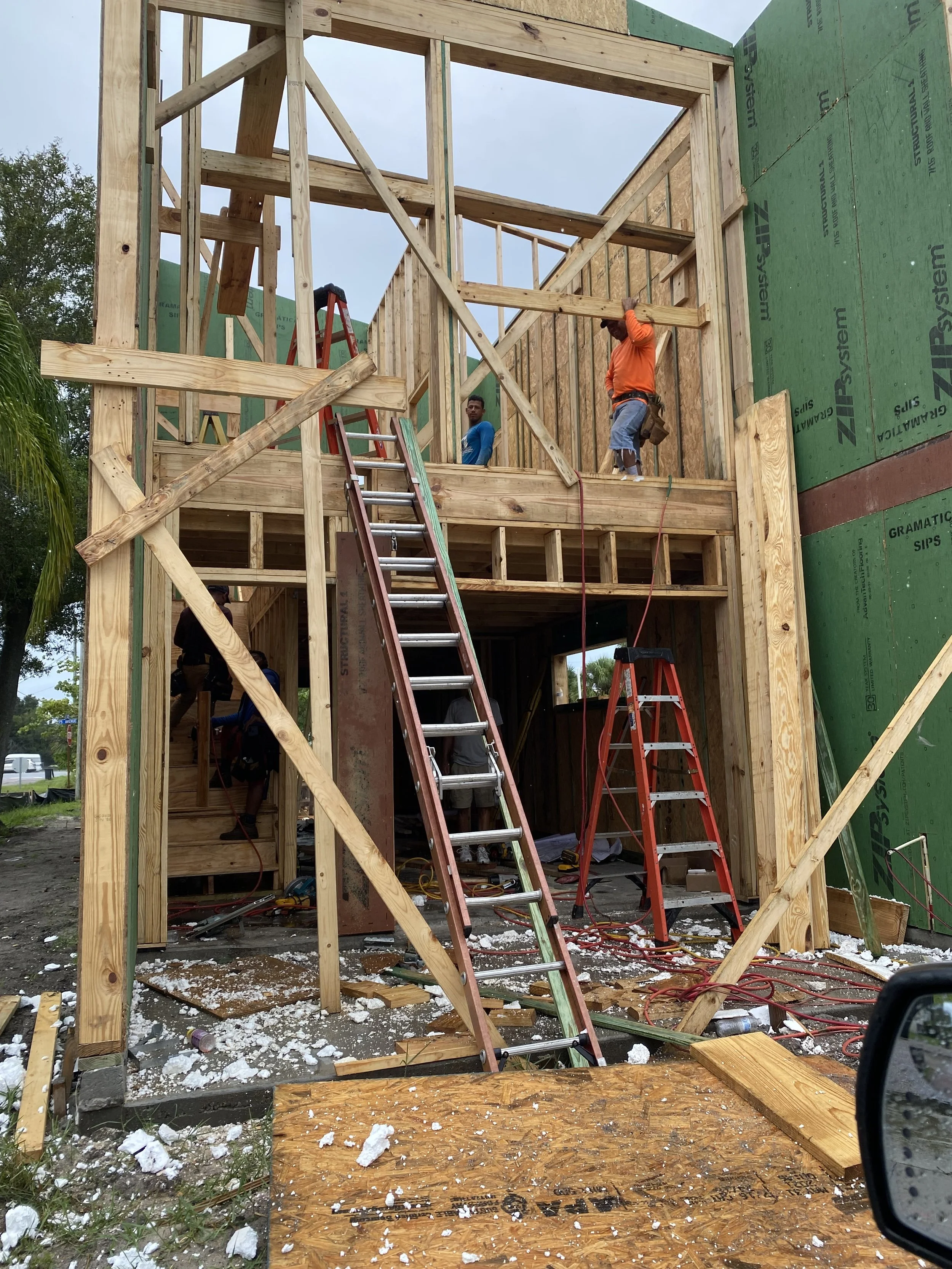 A house under construction with wooden framing, ladders, and construction workers working on the upper level. Debris and insulation material scattered on the ground.