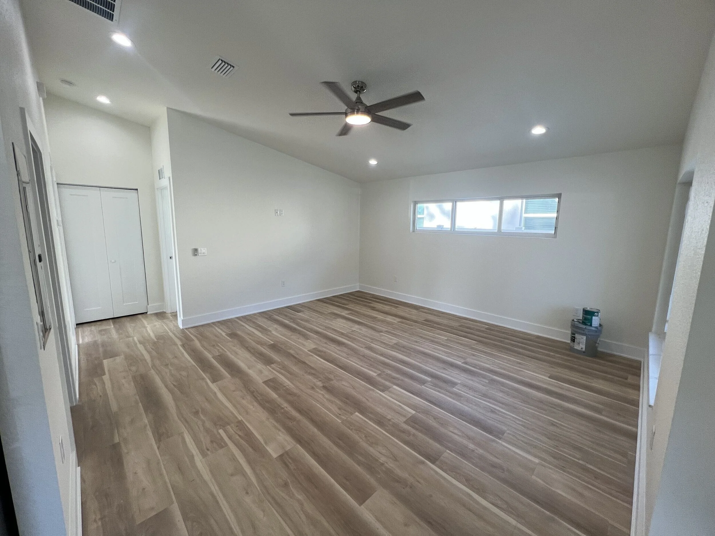 Empty living room with hardwood floors, white walls, a ceiling fan with light, and a window.
