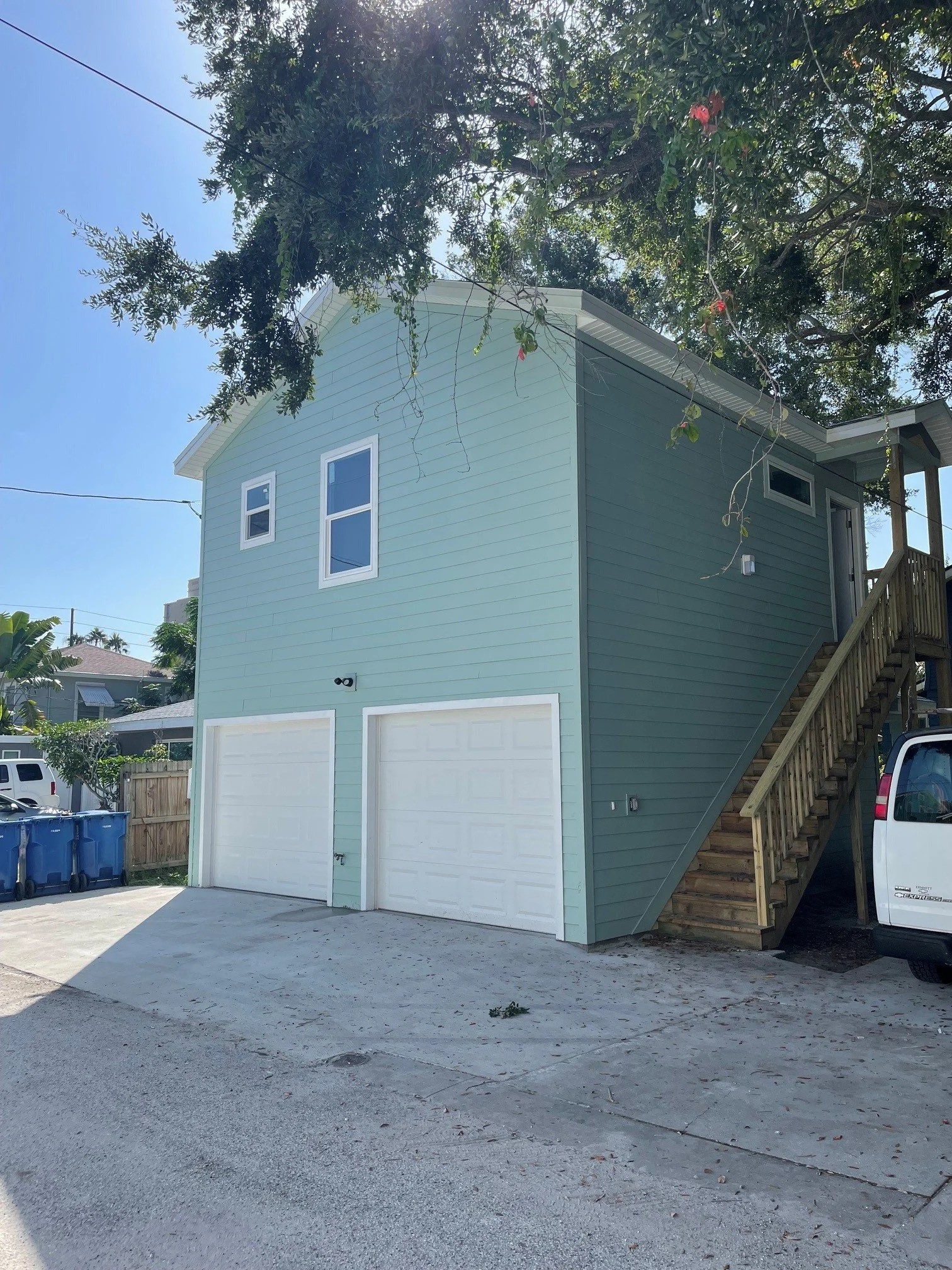 A two-story house with green siding, white trim, and two white garage doors. Exterior stairs lead to an entrance on the second floor. There are trees, a wooden fence, and some blue recycle bins in the driveway. The sky is clear and sunny.