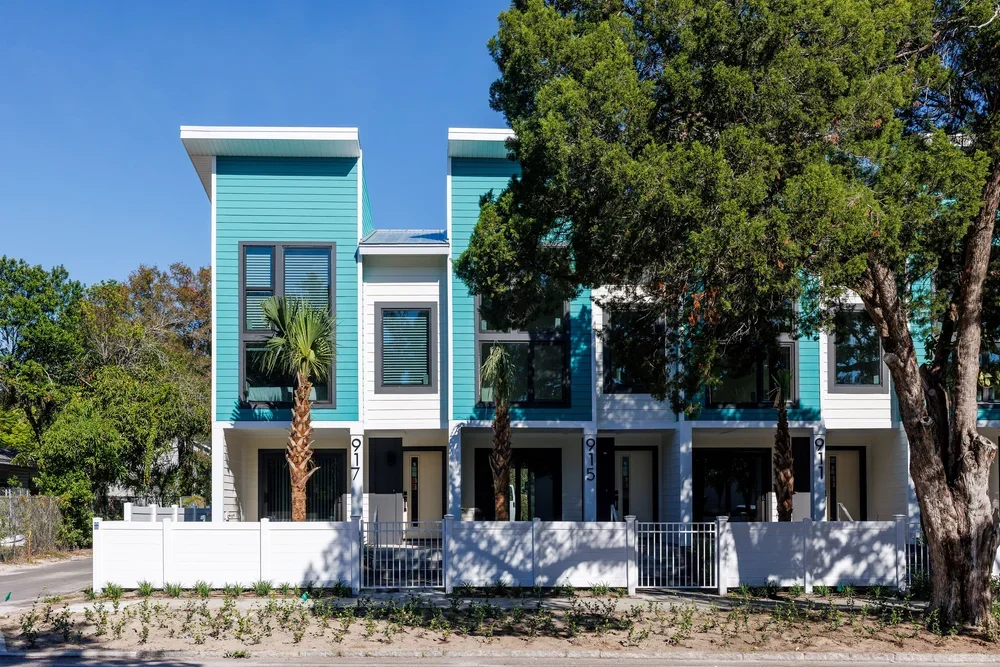 Modern two-story townhouse with blue siding, white trim, large windows, and small front yard gated with white fence.