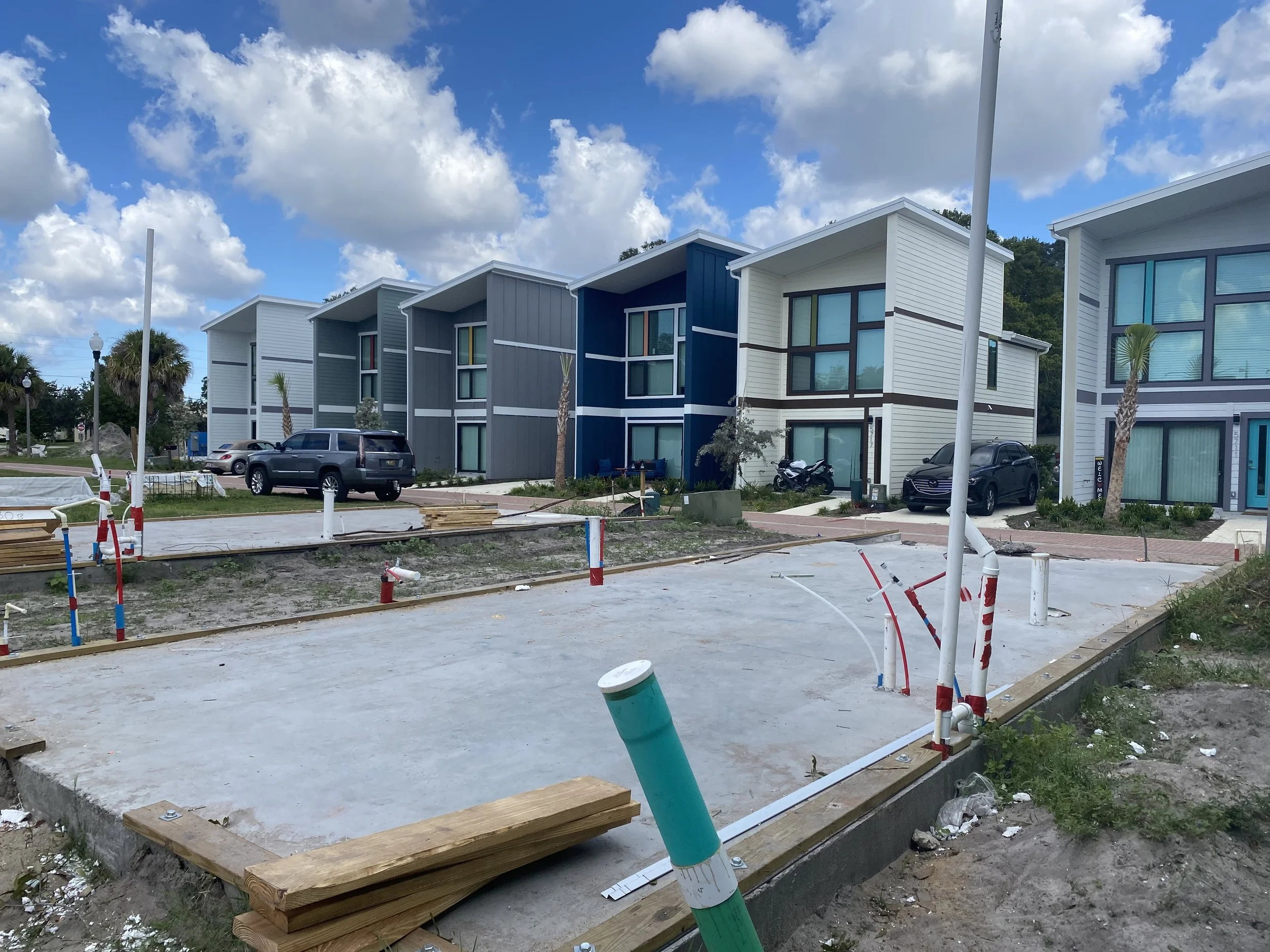 Construction site with a concrete foundation, pipes, and wooden planks in front of modern multi-story houses with large windows and colorful exterior panels, under a partly cloudy sky.
