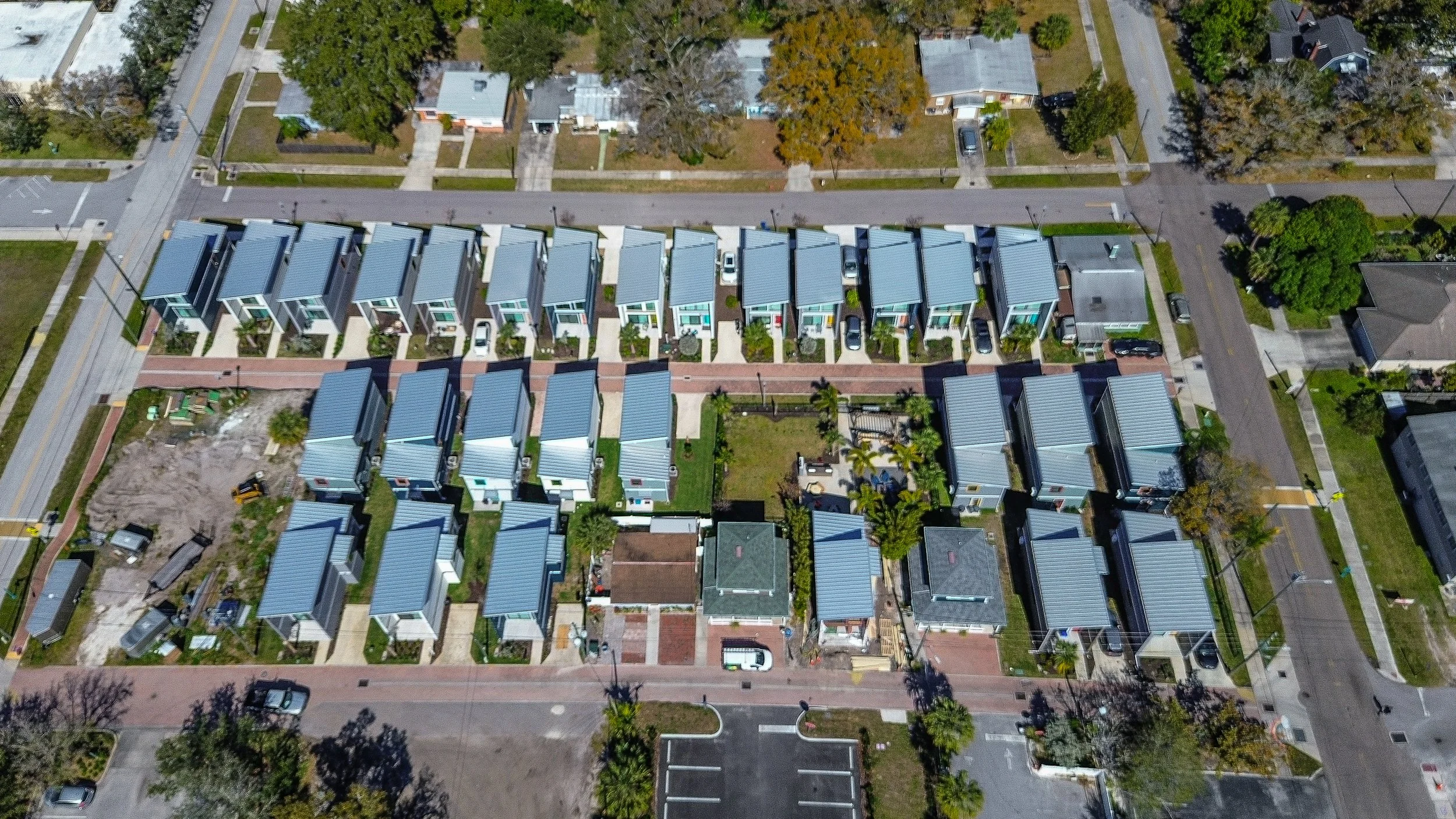 Aerial view of a neighborhood with rows of modern houses with blue roofs, parked cars, and trees lining the streets.