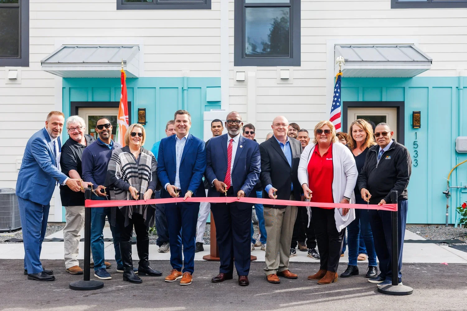 A group of people at a ribbon-cutting ceremony in front of a new building with a light blue door and white siding. The group includes men and women dressed in business and casual attire, standing behind a red ribbon with scissors in hand.