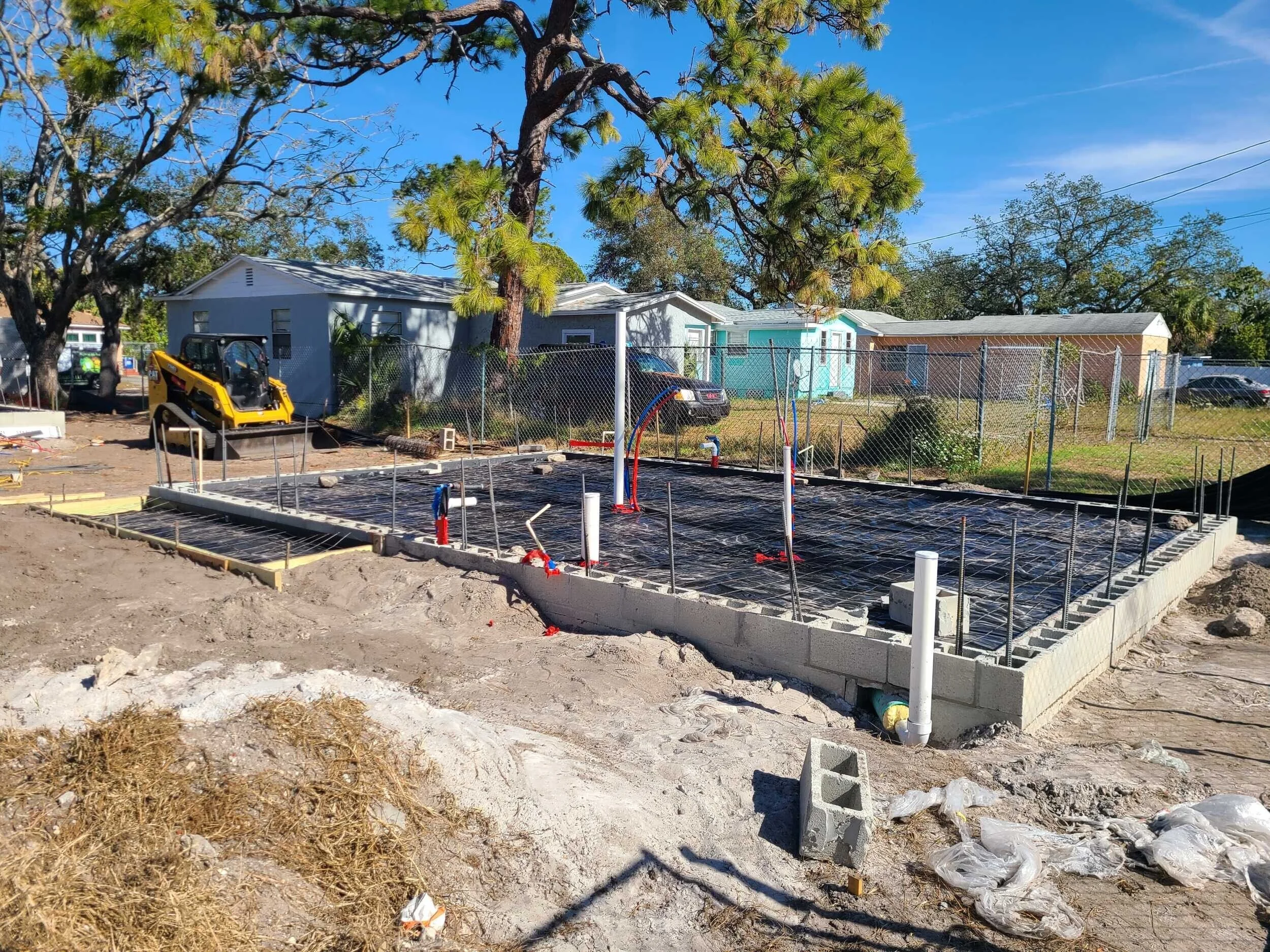 Construction site with foundation being built, cinder blocks, pipes, and a small yellow construction vehicle in front, behind a chain-link fence, with residential houses and trees in the background under a clear blue sky.