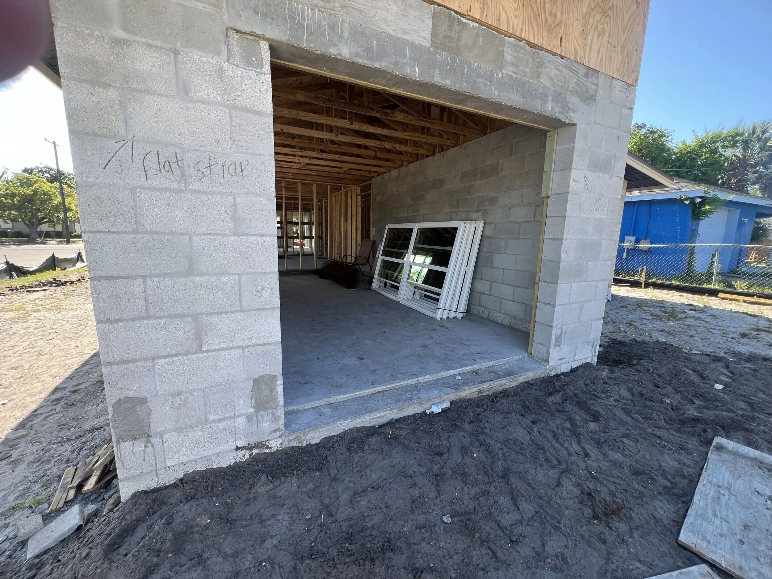 Construction site showing a building under construction with exterior concrete block walls, a wooden roof structure, and window frames leaning against the wall.