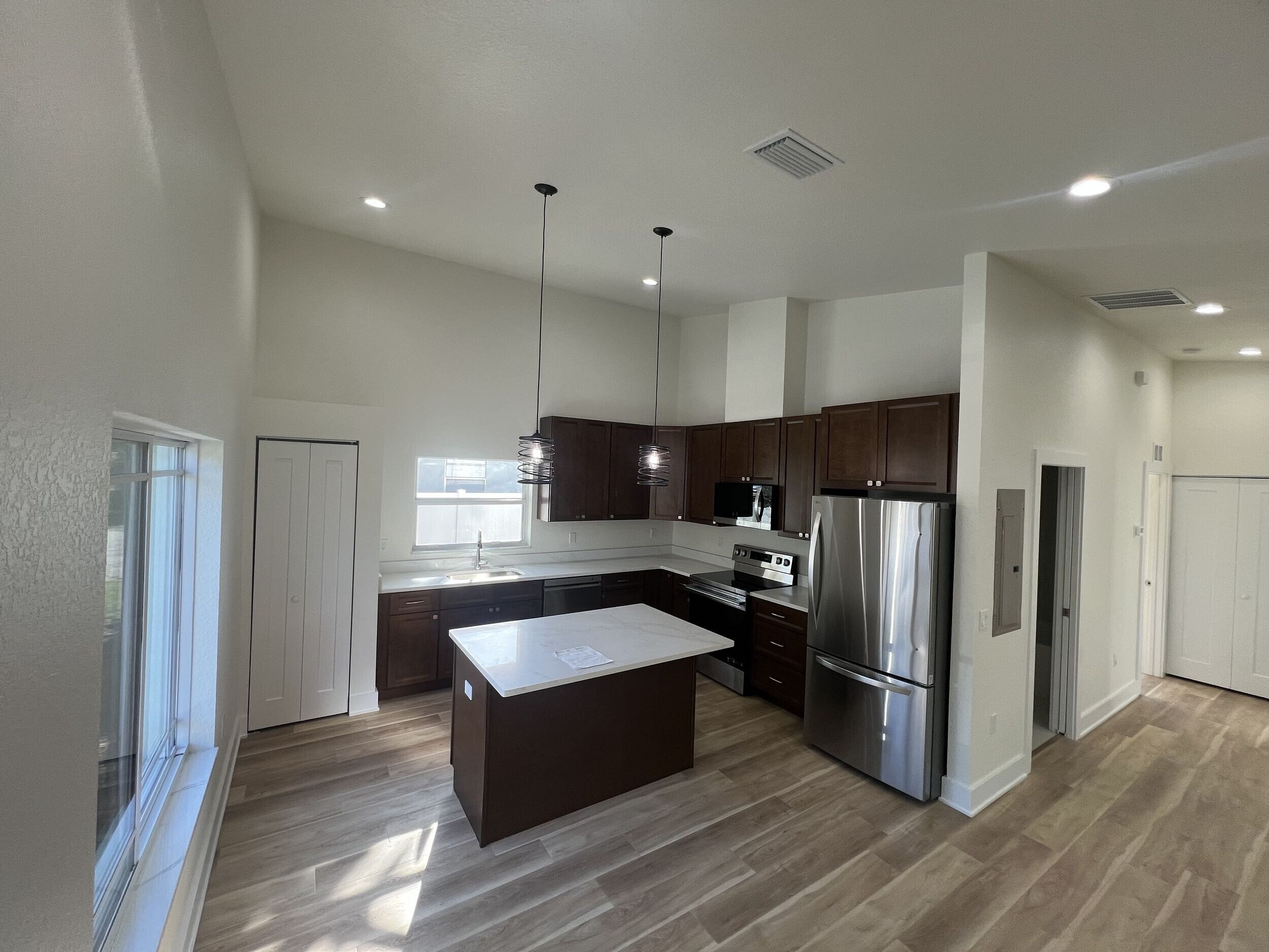 Modern kitchen with dark wood cabinets, stainless steel appliances, island center, pendant lights, large windows, and wood-look flooring.