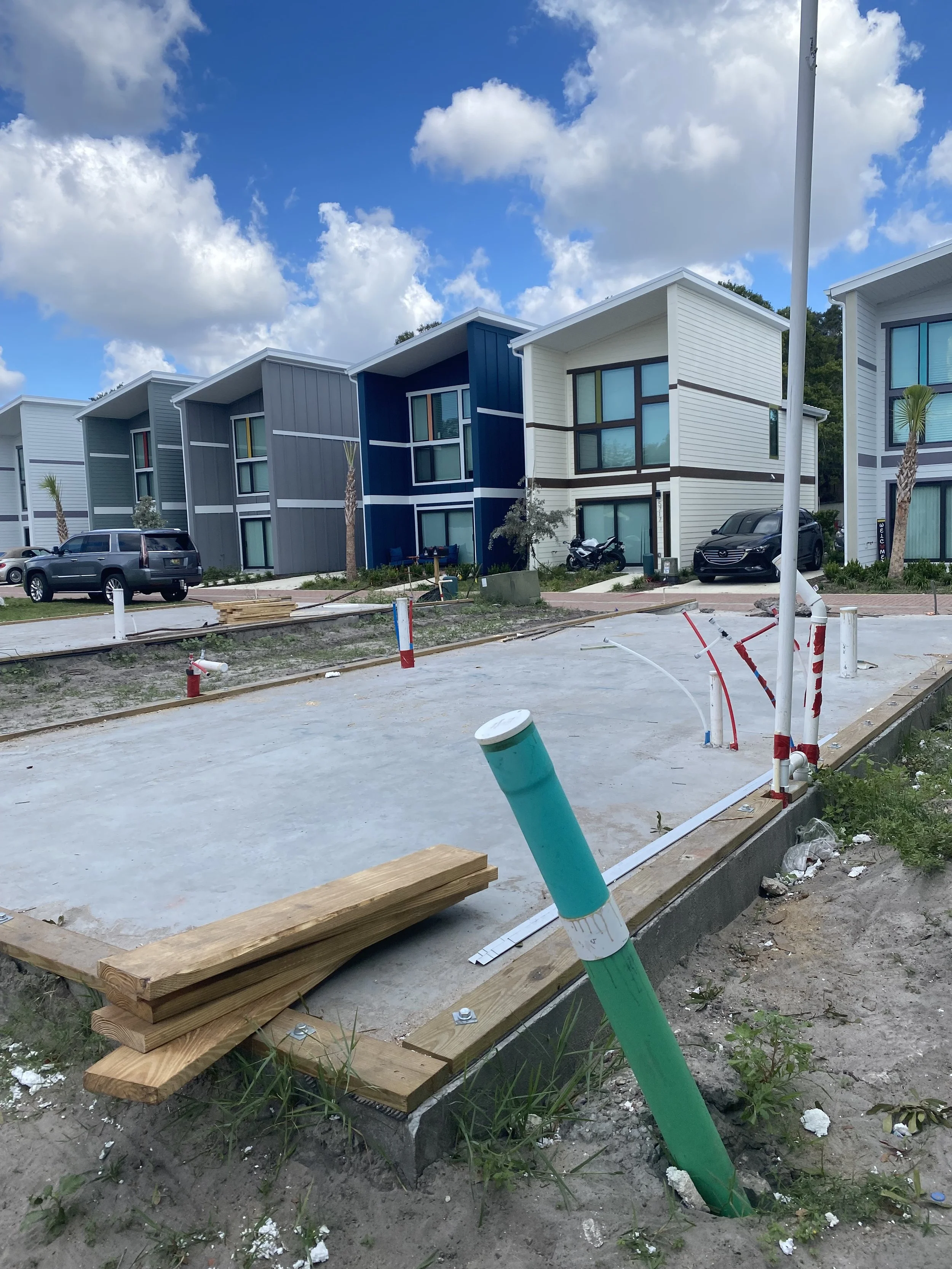 Construction site with colorful modern houses, vehicles parked, partially built sidewalk with exposed pipes and wooden planks in the foreground, under a partly cloudy sky.