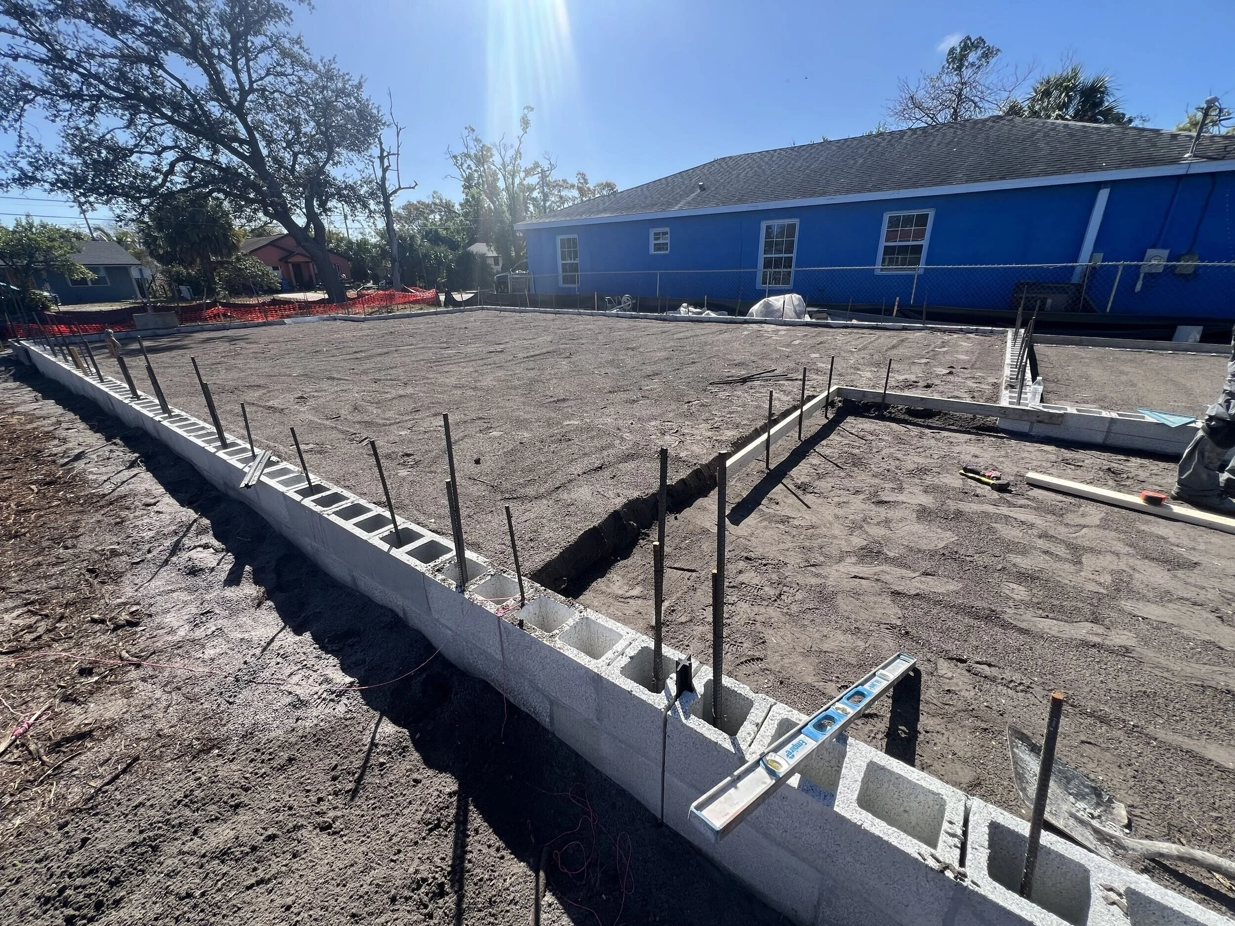 Construction site with concrete foundation and framing for a building, surrounded by trees and residential houses.