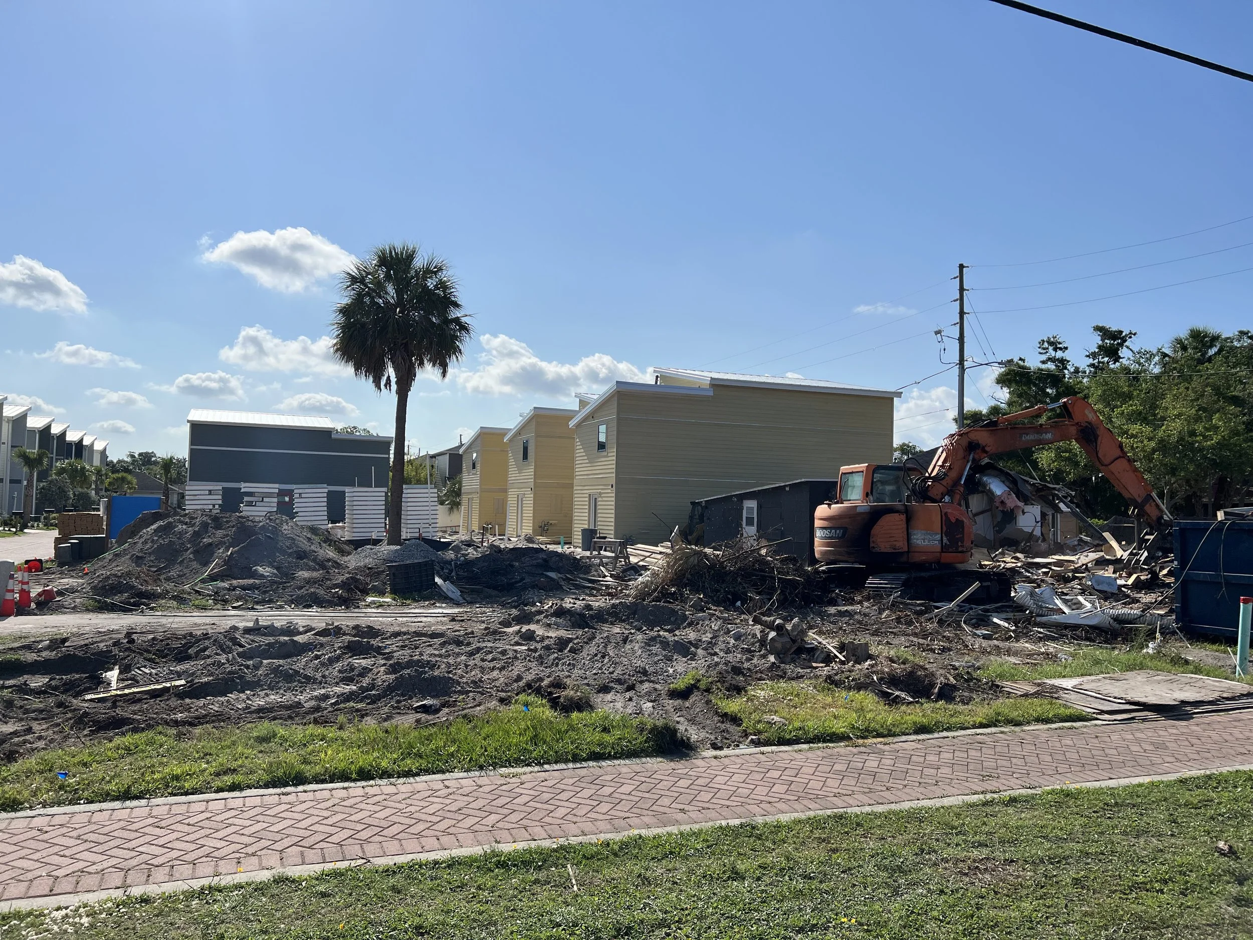 A construction site with excavated dirt, construction debris, a small orange excavator, and residential buildings in the background under a bright blue sky with scattered clouds.