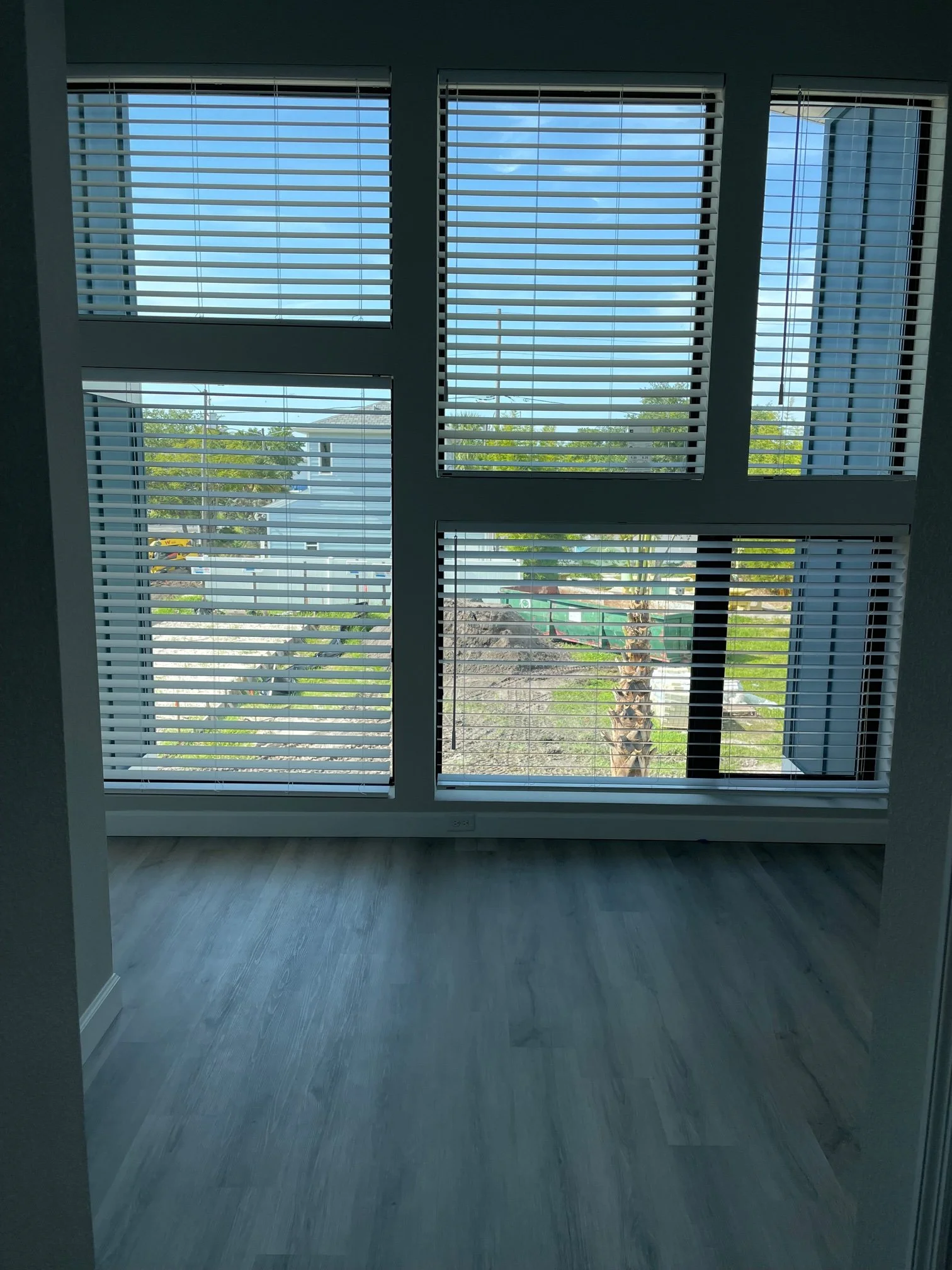 Interior view of a room with large windows and blinds, showing a street, trees, and neighboring houses outside.