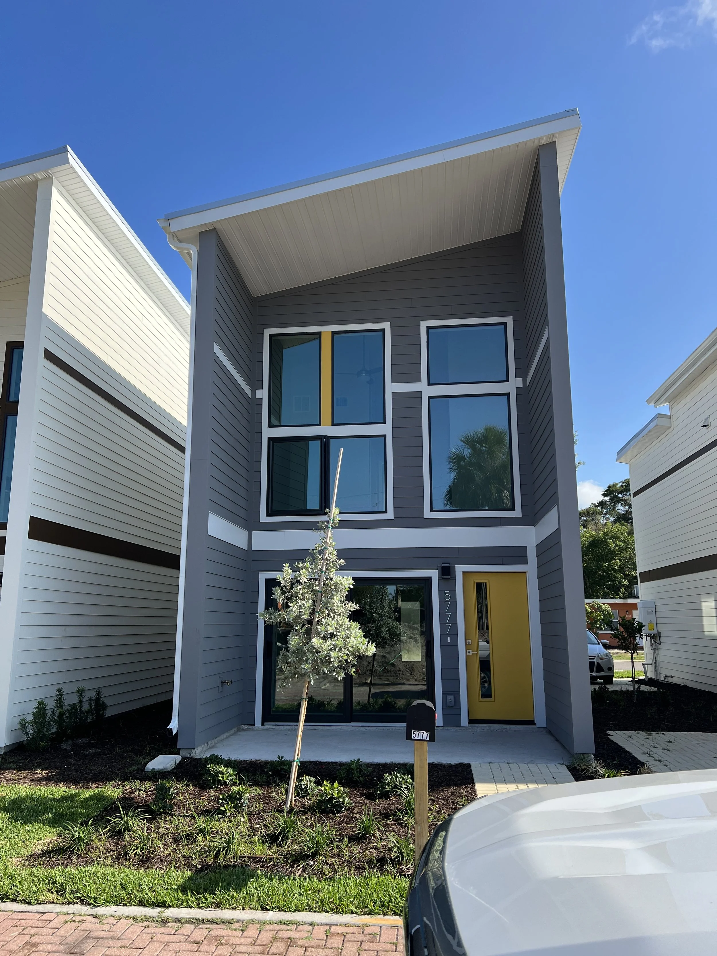 A modern multi-story house with a gray exterior, large windows, and a yellow front door. There is a small tree and landscaping in front, a mailbox with the number 5777, and a white car in the foreground. The sky is clear and blue.