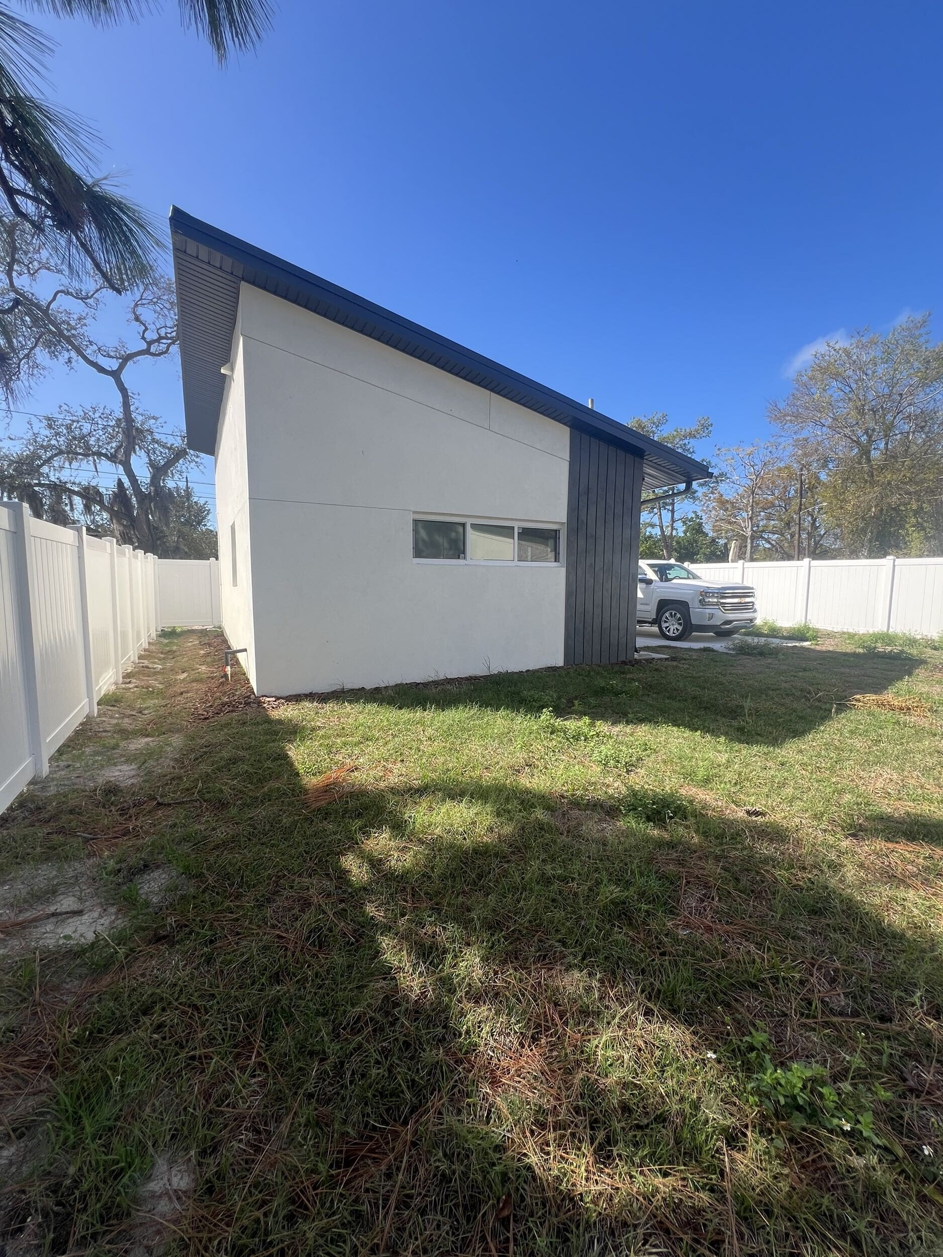 A modern house with a dark blue roof and white exterior walls, with a horizontal window, is visible behind a white fence. A white pickup truck is parked in the driveway on the right side. The sky is clear and blue, and there are some trees with sparse leaves in the background. The foreground consists of grass and patches of dirt.