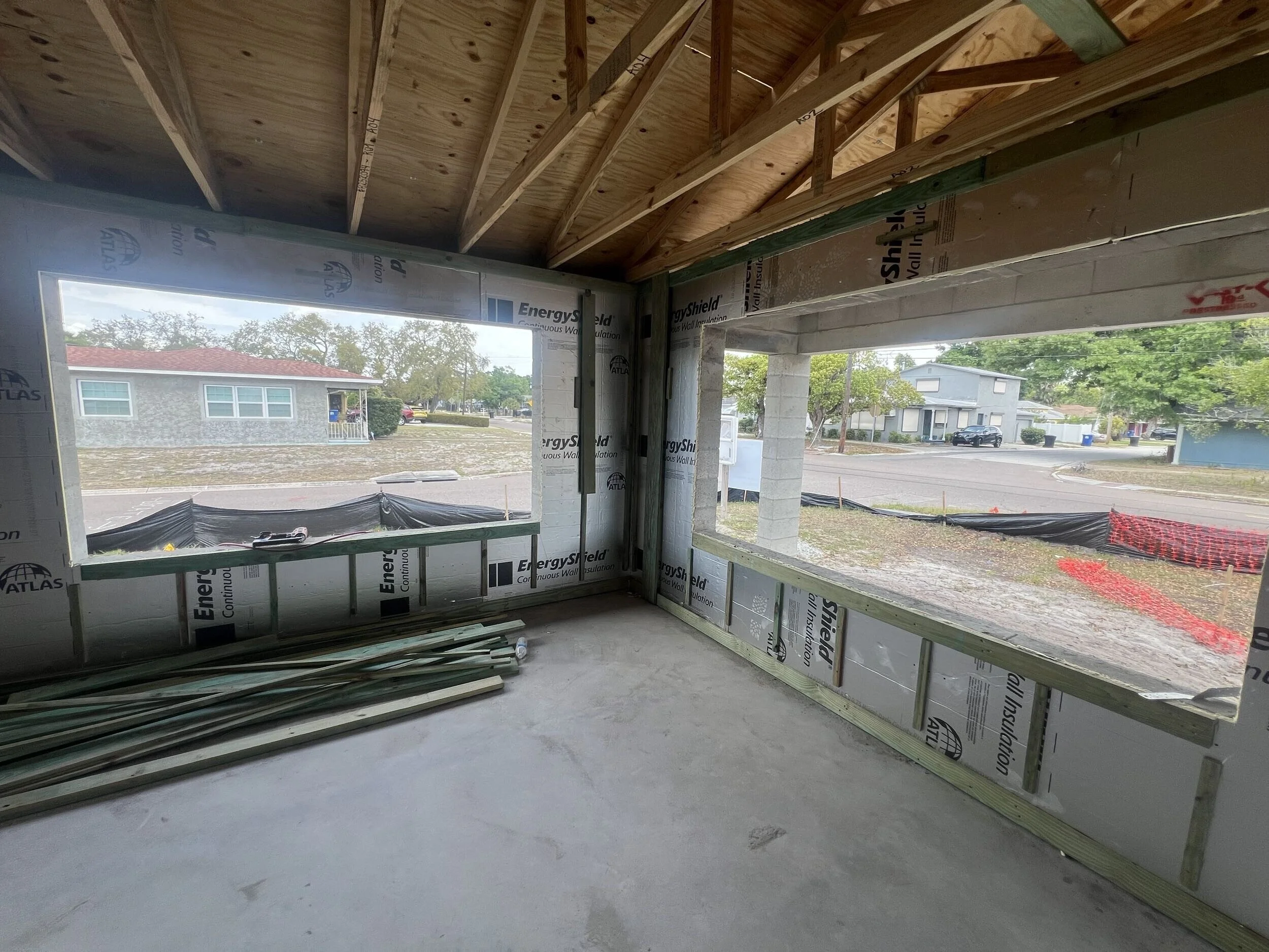 Interior view of a house under construction showing an unfinished room with large window openings, construction materials and wall insulation with brand labels, and visible wooden framing and roof support beams