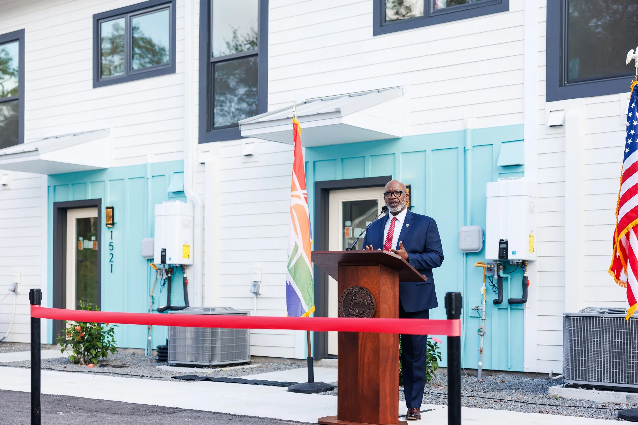 A man in a suit speaking at a podium outdoors in front of a new building with construction elements visible. He's wearing glasses and a red and white striped tie, with an American flag and a rainbow pride flag beside him.