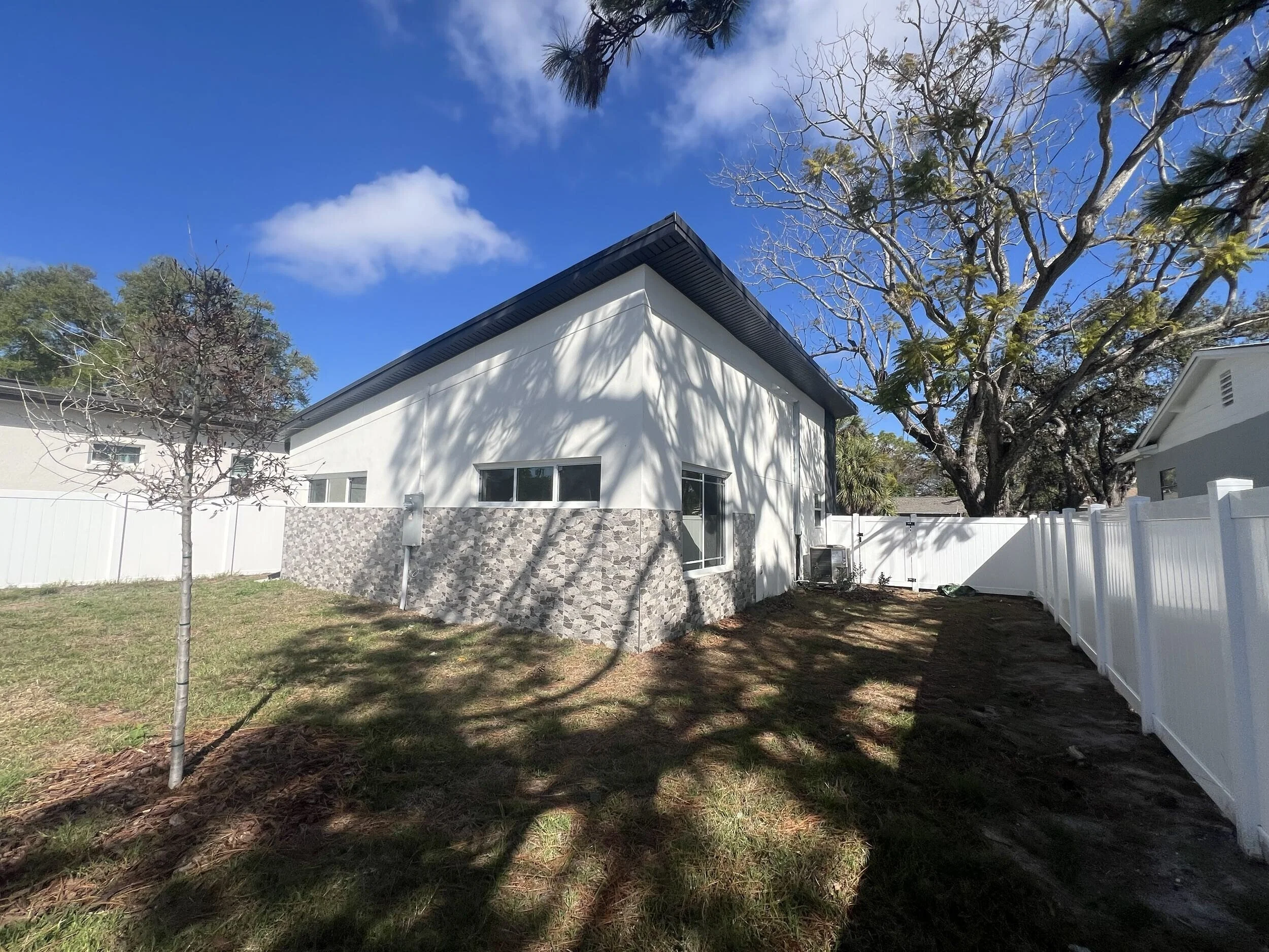 A white house with a black roof, a stone wall on one side, and windows on the front, surrounded by a white fence, trees, and grass, under a blue sky with a few clouds.