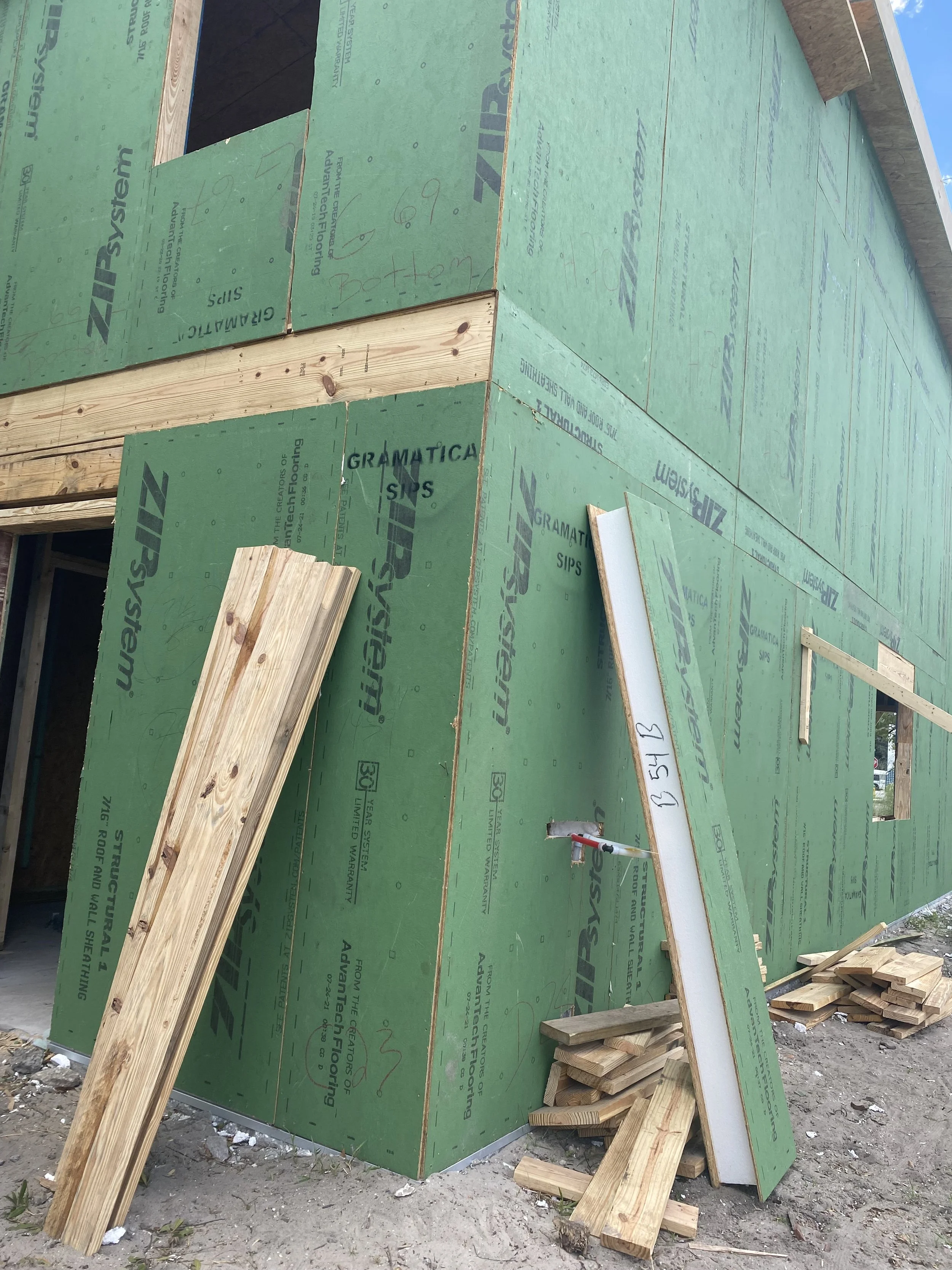 Construction site with partially built wall covered in green insulation material, wooden panels leaning against it, and a small pile of wood pieces on the ground.