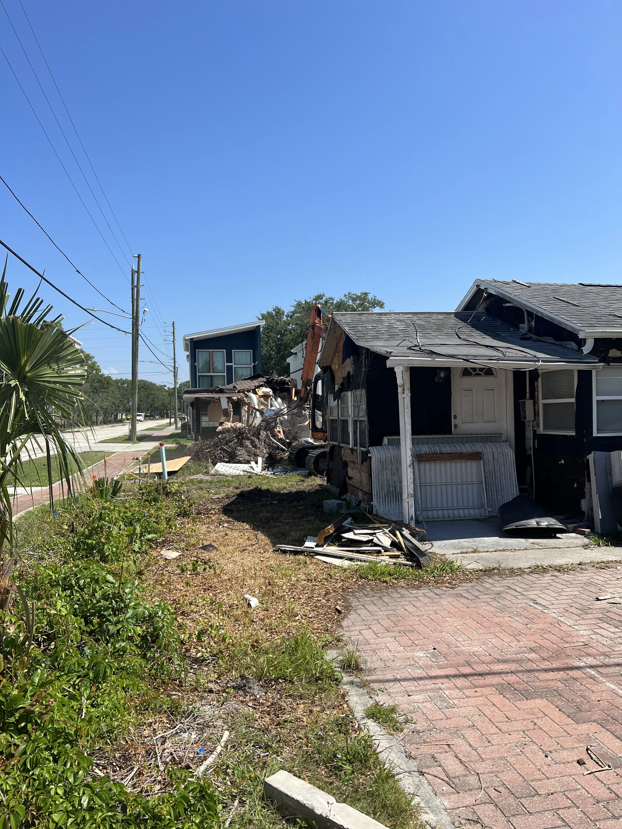 Residential house with extensive damage from an apparent explosion or fire. Roof and walls are severely damaged, debris scattered around. Clear blue sky and power lines in the background.