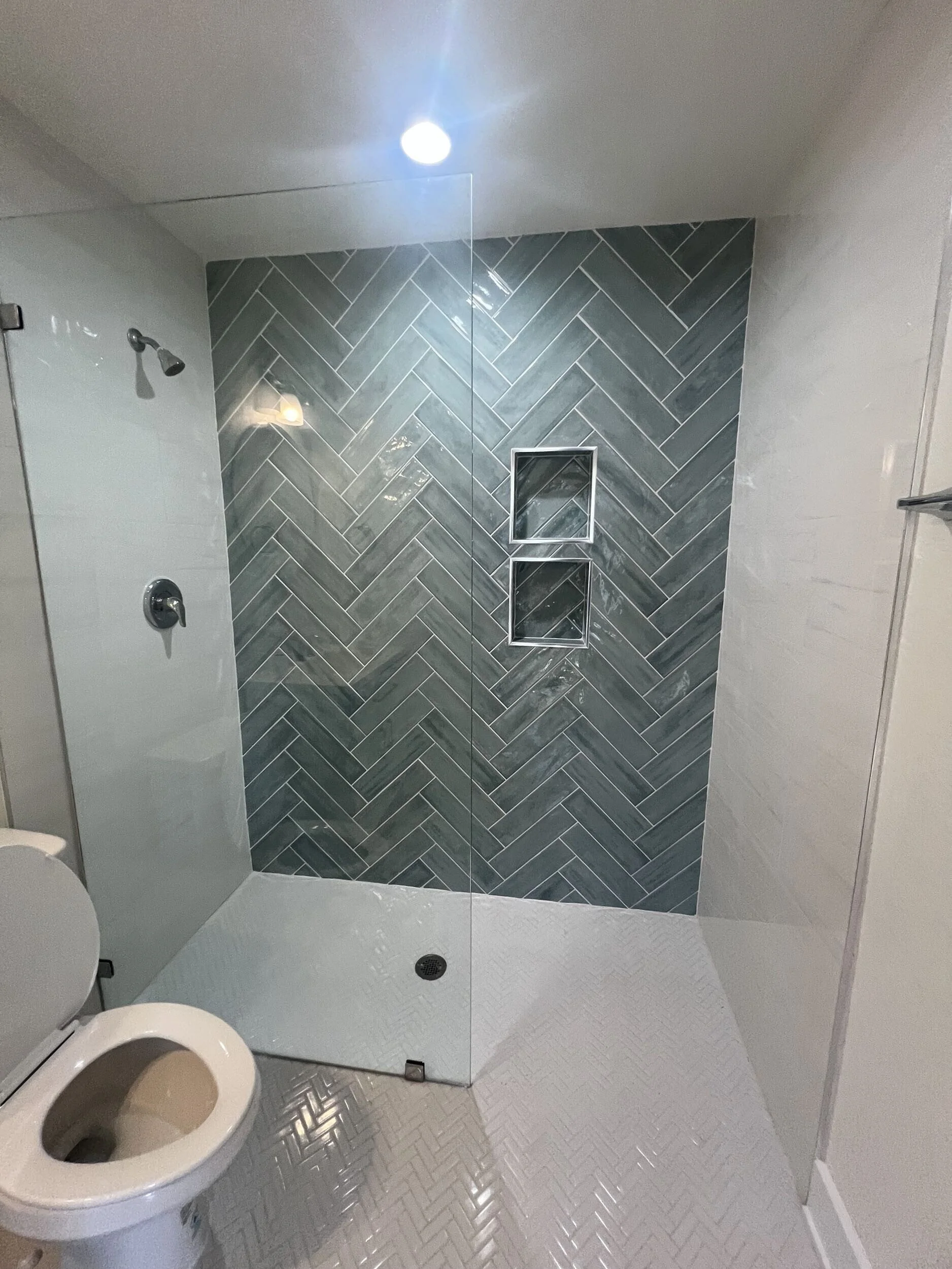 A bathroom with a glass shower enclosure, a tiled shower wall in a herringbone pattern, and built-in shelves. The bathroom has a white toilet and a white floor with a textured pattern. There's a ceiling spotlight providing illumination.