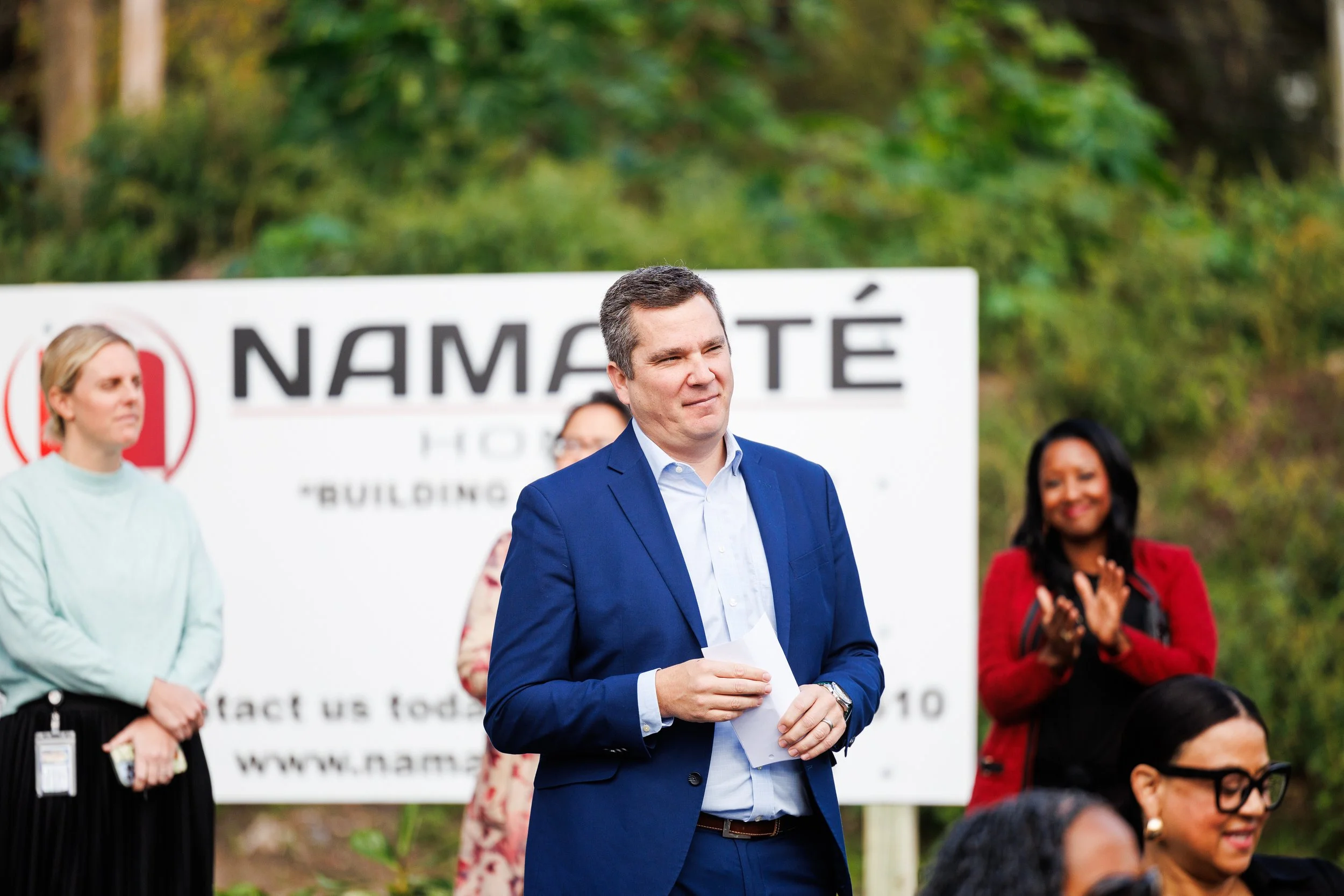 Man in blue suit speaking at outdoor event with women clapping and listening, behind a sign that reads 'NAMASTE'.