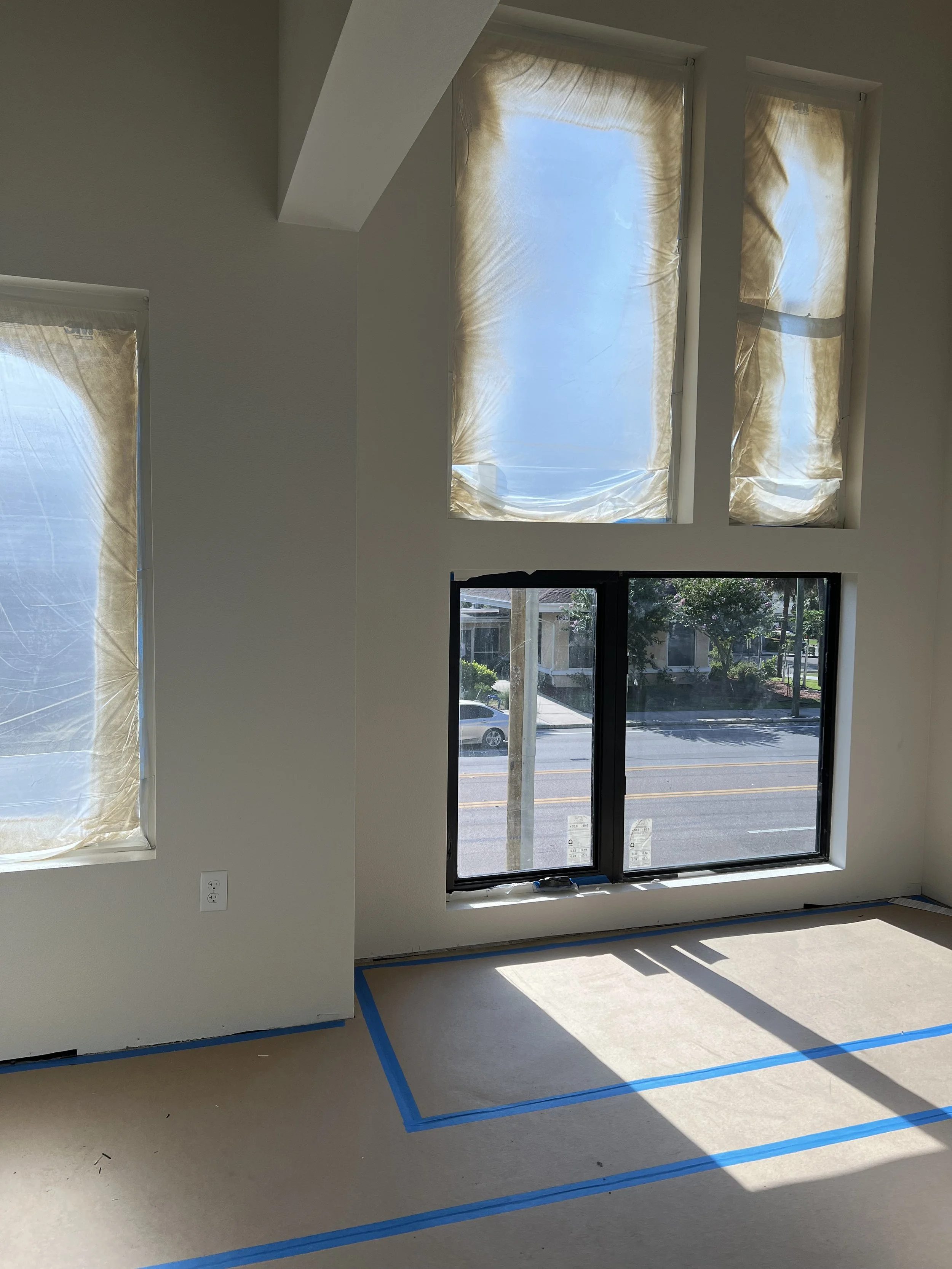 Interior of a room under construction with large windows covered with plastic sheets, taped edges on the floor, and two electrical outlets on the wall.