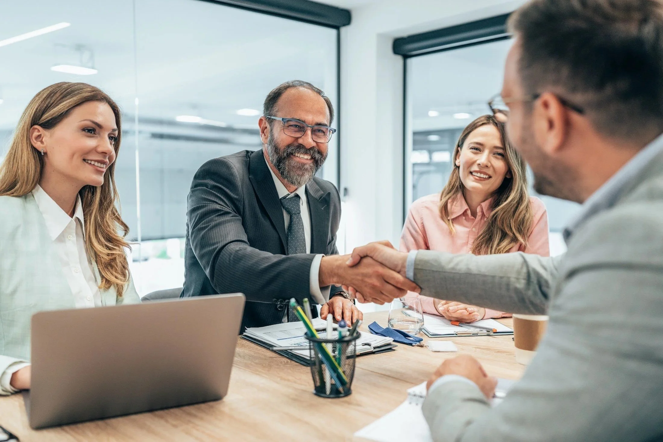 A group of five business professionals in a meeting room, with one man shaking hands with another. They are smiling and appear to be engaged in a discussion. On the table, there are notebooks, pens, a laptop, and coffee cups.
