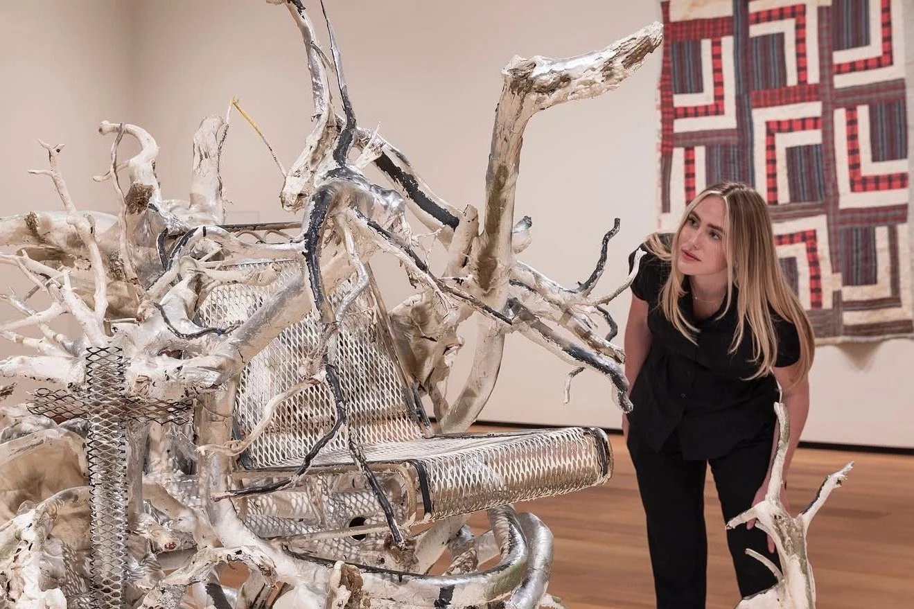 A woman in black clothing carefully observes a contemporary art sculpture made of intertwined, weathered tree branches and metal mesh, displayed in an art gallery with a quilted wall hanging in the background.