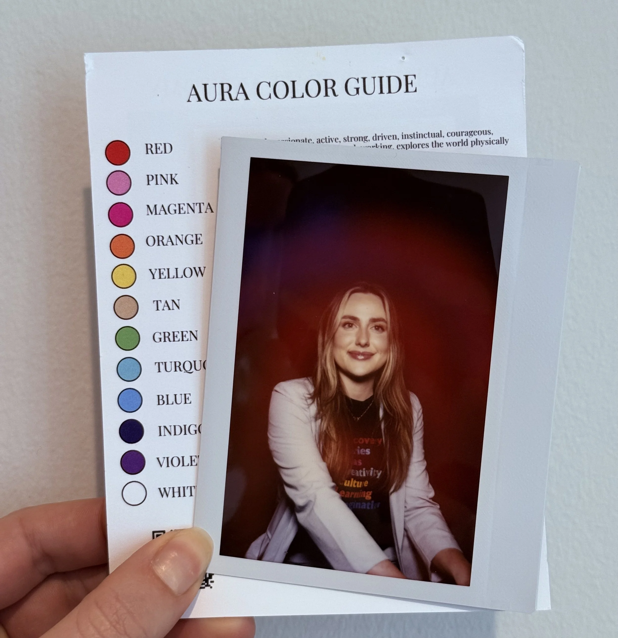 Close-up of a color guide card with the title "AURA COLOR GUIDE" and a list of colors including red, pink, magenta, orange, yellow, green, turquoise, blue, indigo, violet, and white on the left side. A person's hand is holding the card, and there's a blurred photograph of a woman with long hair and a white blouse in the background.